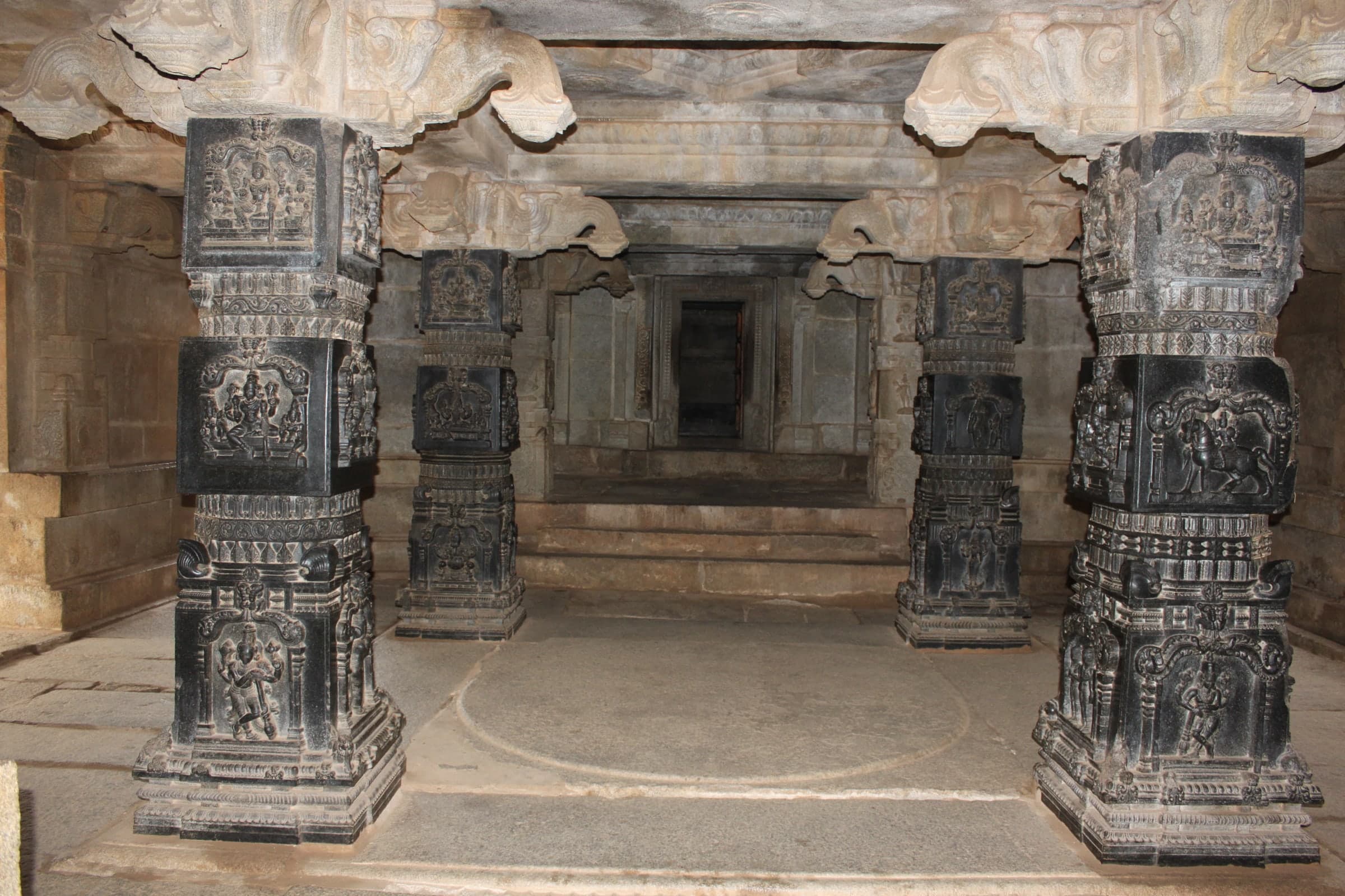 Four dark, densely carved stone pillars in a hall, two in the foreground and two behind, with carved figures in panels on each face. Beyond them, steps and a series of doorways lead to a dark inner chamber.
