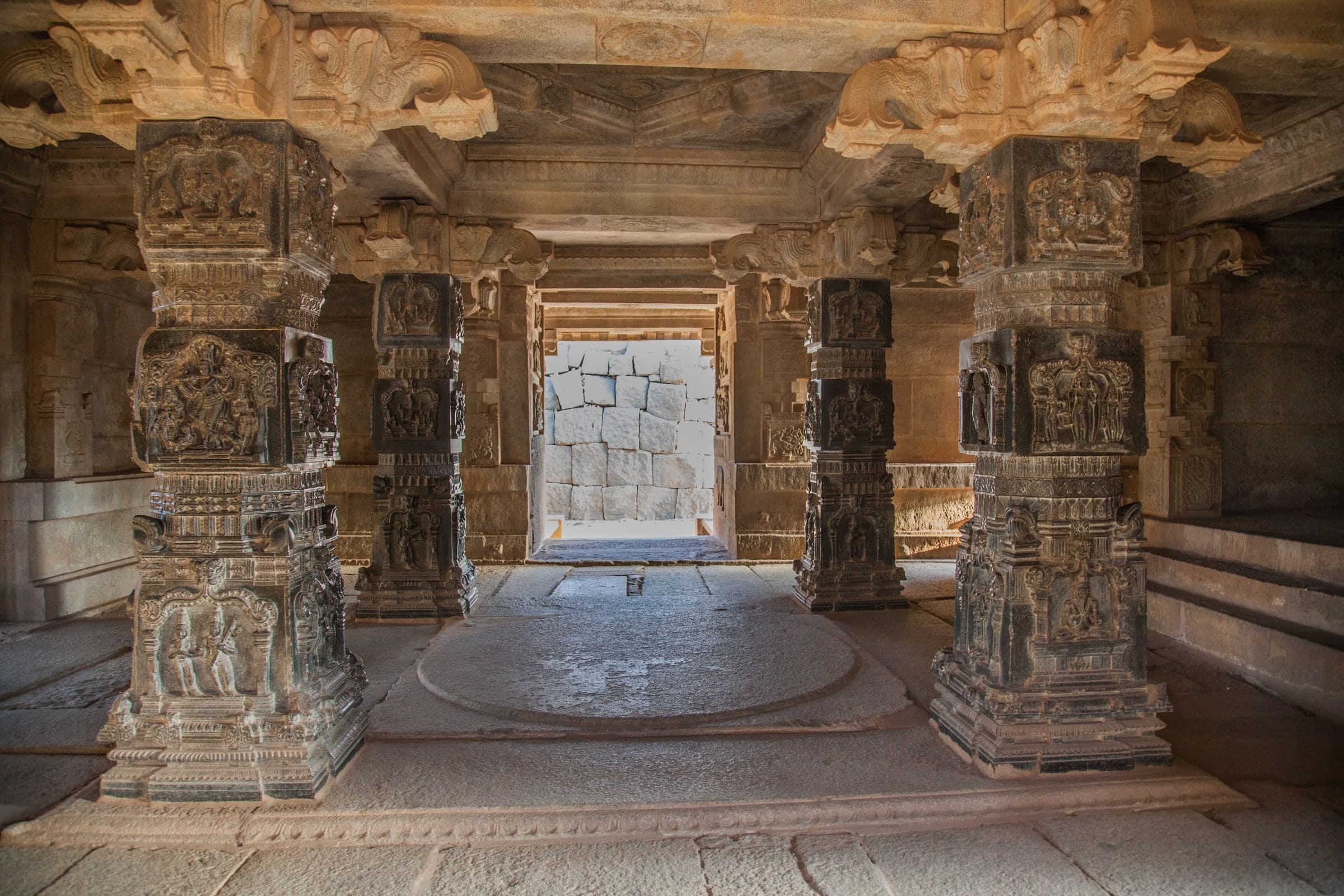A dim stone hall with four square pillars of dark polished stone, each carved in stacked panels of figures, standing around a raised circular floor slab. A doorway at the far end opens onto sunlit stone blocks.