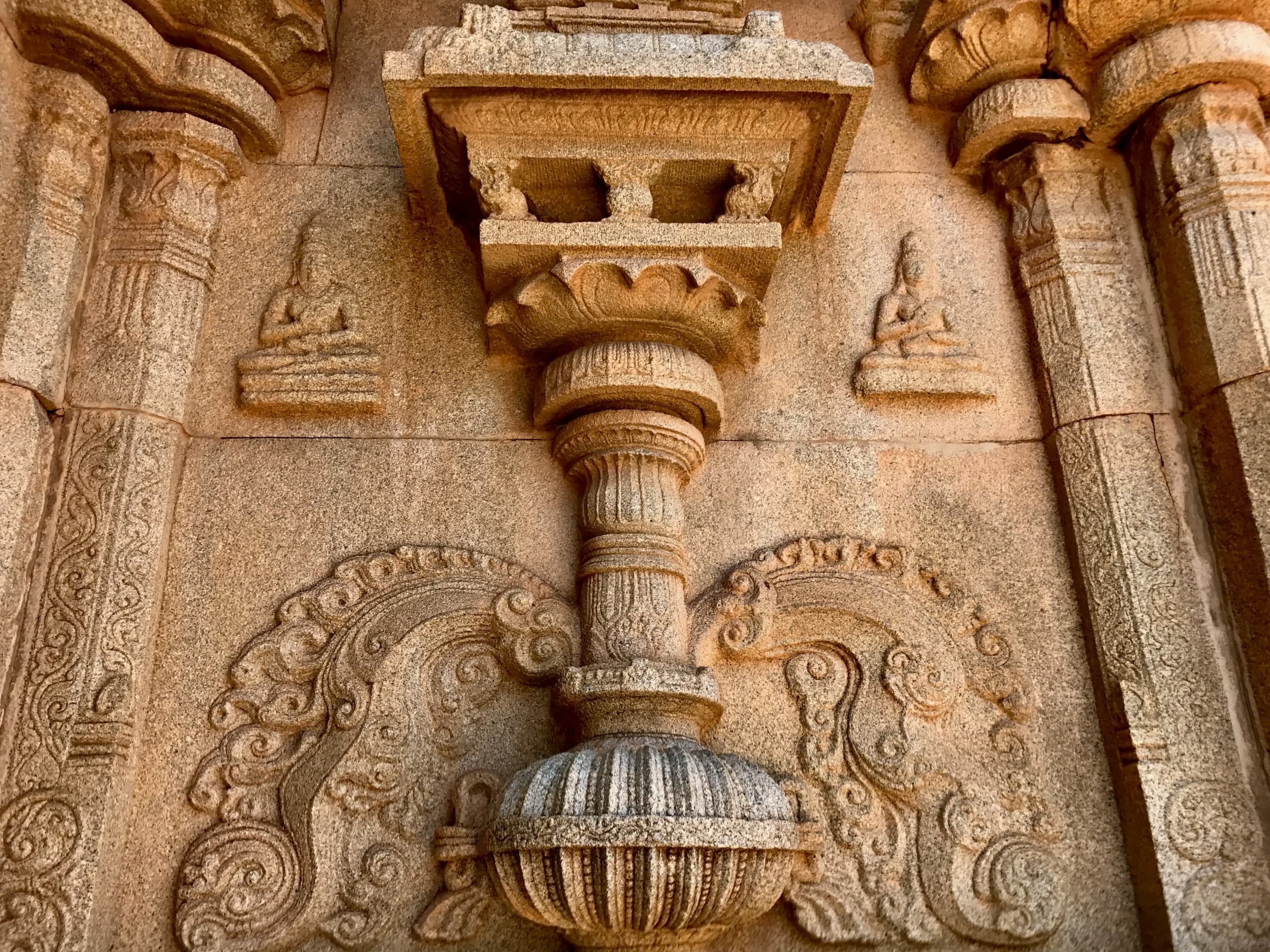 A granite wall with a carved pilaster rising from an ornate pot base, framed by scrolling foliage. On either side, high on the wall, a small figure sits cross-legged in meditation on a pedestal.