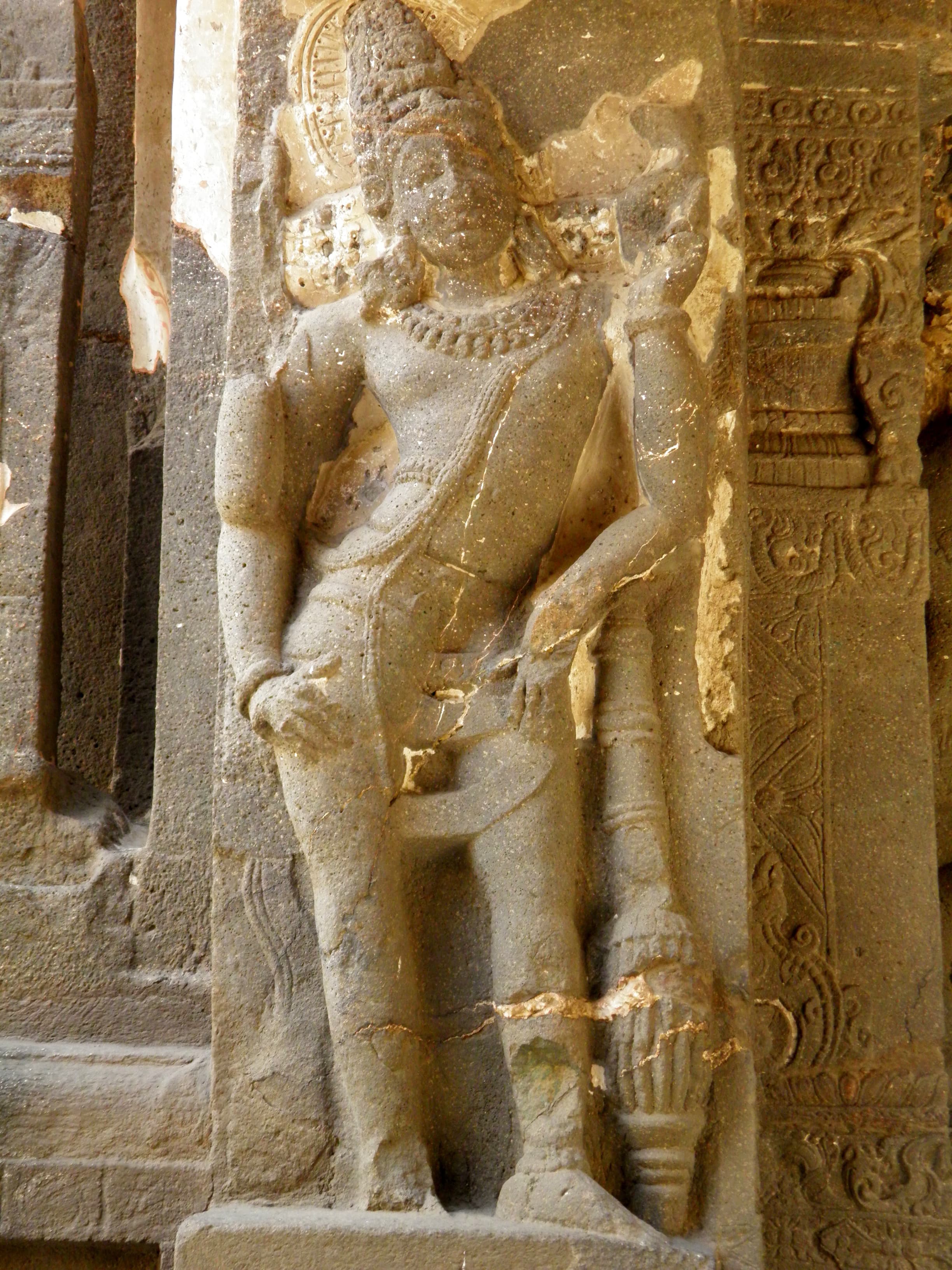 Tall rock-cut dvārapāla at Kailāsa Temple, Ellora Cave 16. The crowned guardian stands in a relaxed posture against a carved pilaster, one hand resting low, a smaller attendant figure beside him, lit warmly by daylight falling across the stone.