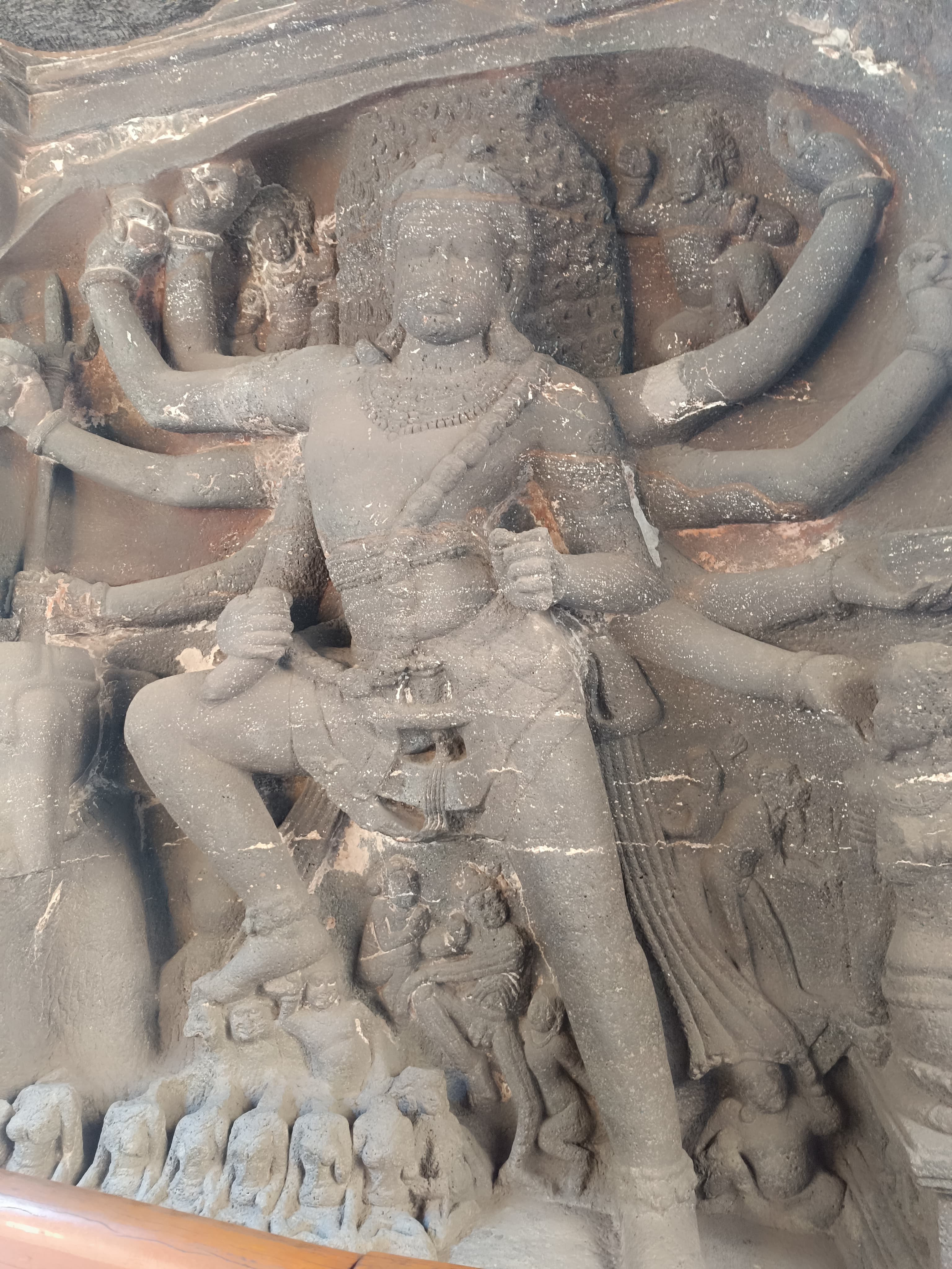 Deep rock-cut relief of a many-armed Śiva striding in a wide, violent stance at the entrance to Kailāsa Temple, Ellora Cave 16. Weapons are held in his outstretched arms, a mass of carved forms rises behind his head, and small attendant figures are crowded in a row along the base of the panel.