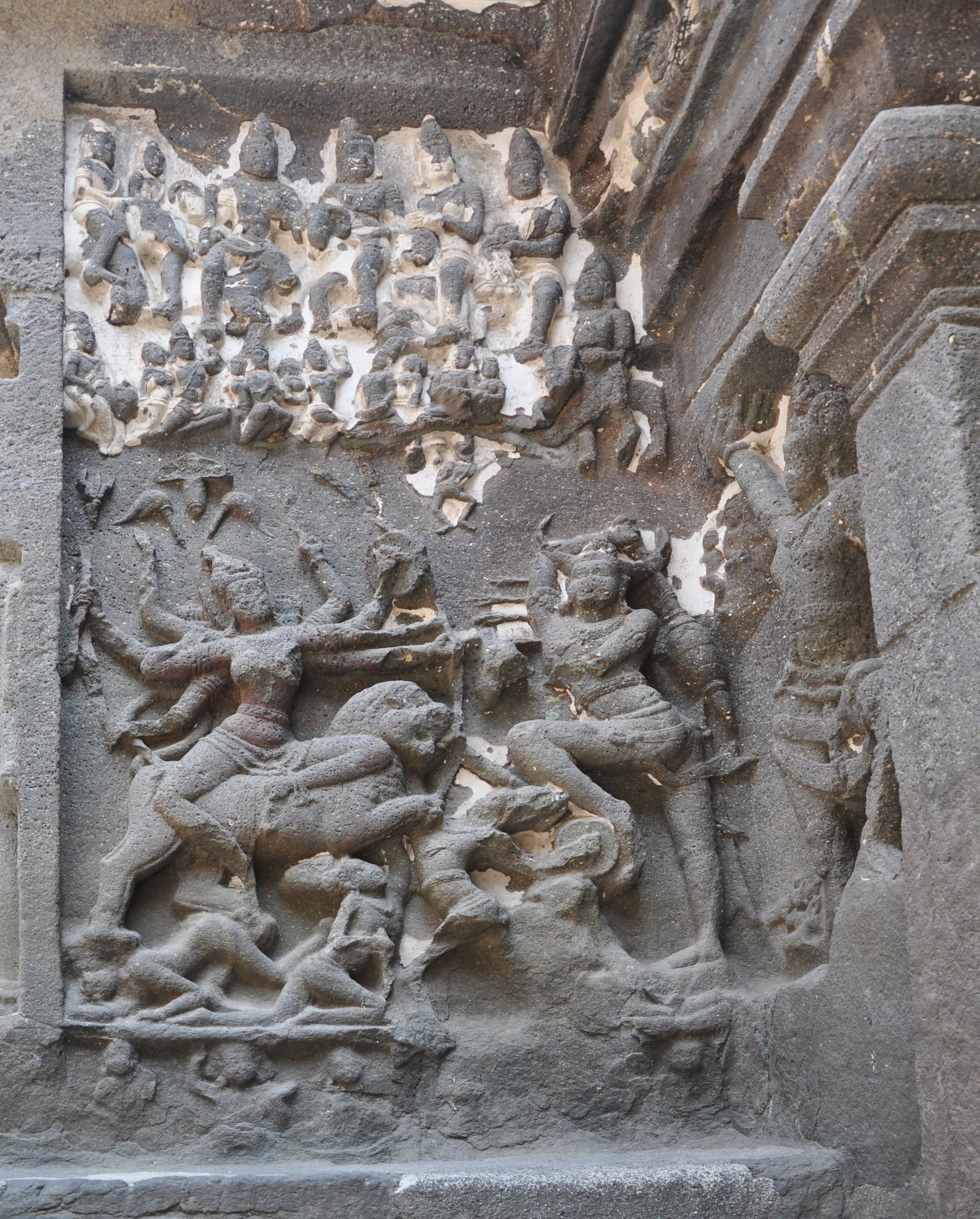 Rock-cut panel at Kailāsa Temple, Ellora. At the left a many-armed Durgā lunges forward with weapons raised; at the right the buffalo-headed demon Mahiṣāsura recoils, his horns clearly carved. A crowded register of small celestial figures fills the upper part of the panel, and fallen figures lie along the base.