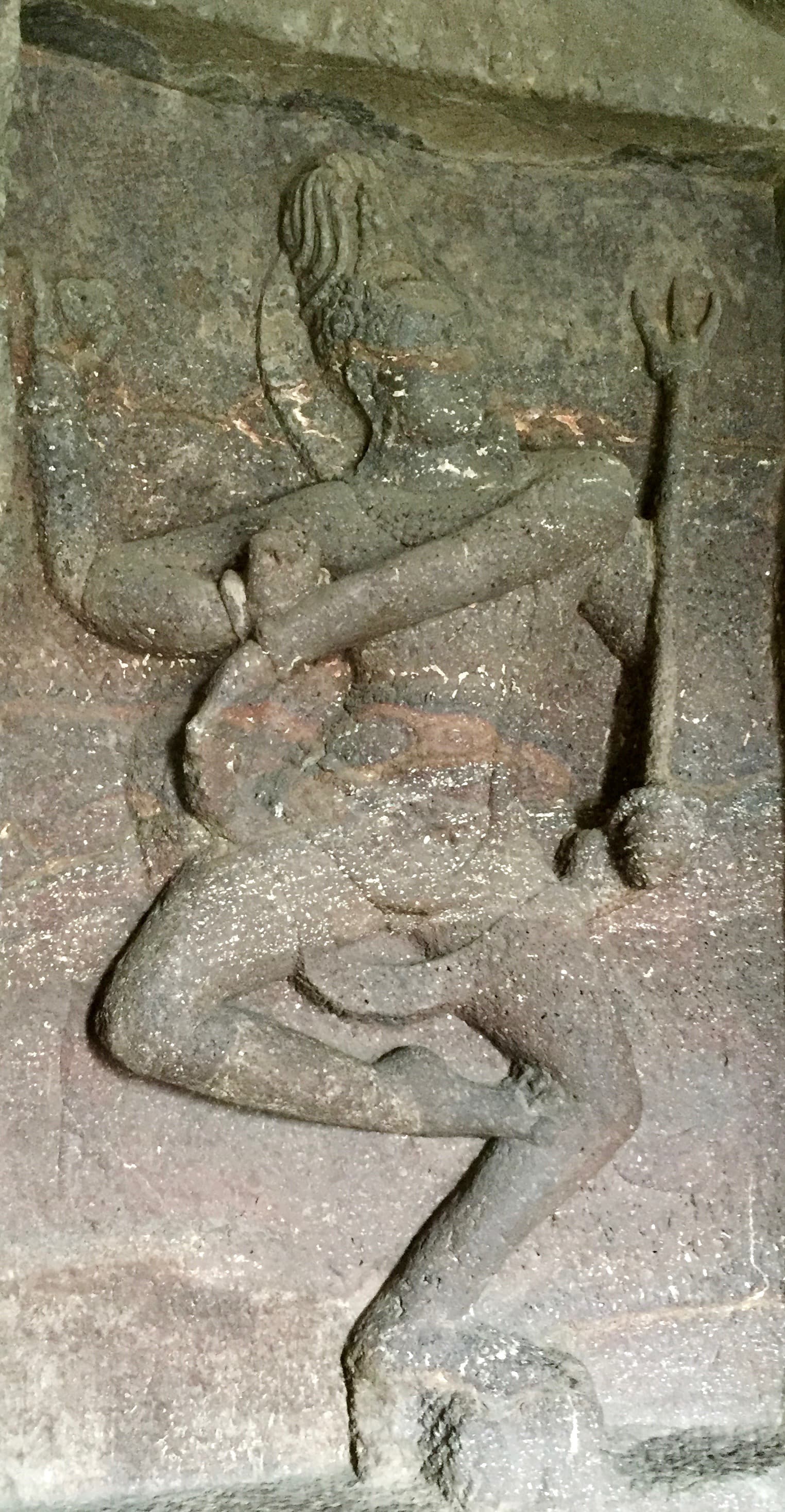 Weathered rock-cut relief of Śiva dancing, carved in a recess at Kailāsa Temple, Ellora. The figure leans into the dance with one leg crossed over the other and a trident standing at the right; much of the surface detail has been lost to erosion.