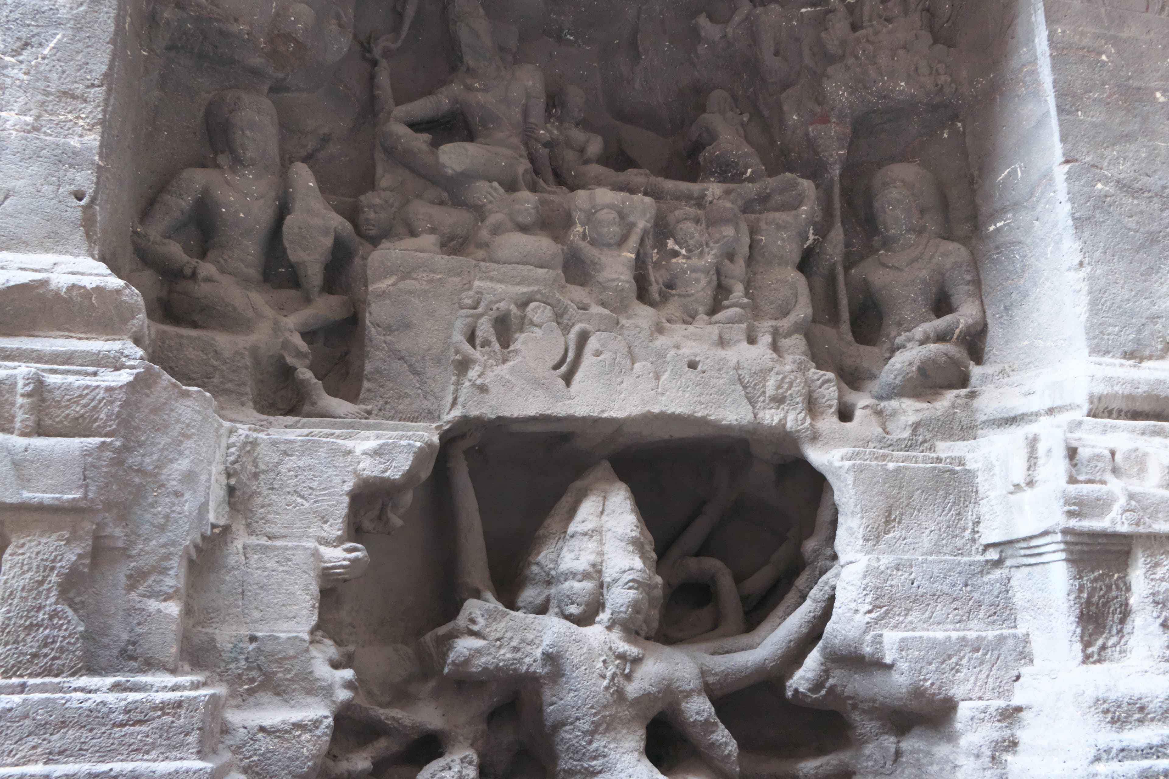 Rock-cut relief at Kailāsa Temple, Ellora. Śiva and Pārvatī sit on a tiered mountain platform with standing attendants and smaller celestial figures around them. In the shadowed recess beneath the platform a many-armed Rāvaṇa crouches, his arms radiating outward and braced up against the underside of the mountain.