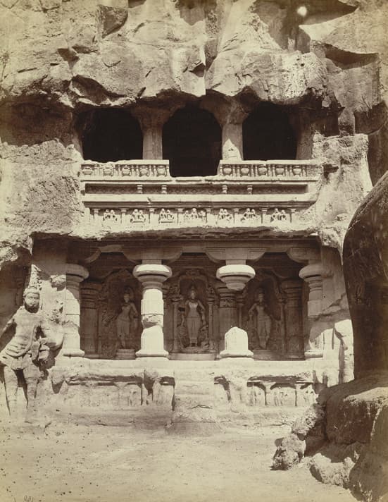 Sepia photograph from 1875 of a two-storey rock-cut shrine cut into the cliff on the north side of the Kailāsa court, Ellora Cave XVI. Three standing river-goddess figures — Gaṅgā, Yamunā and Sarasvatī — occupy separate niches between pillars along the lower storey, with an open pillared gallery above and rough-cut rock overhead.