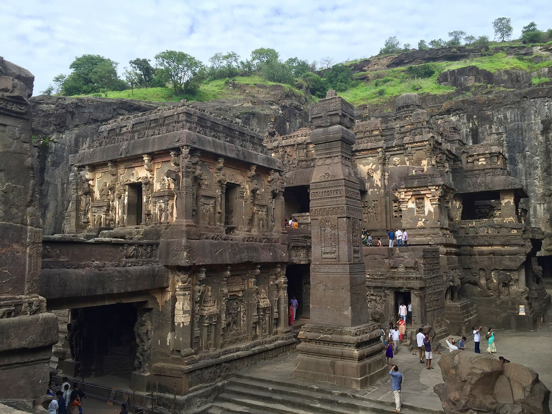 Elevated view within the excavated court of Kailāsa Temple at Ellora. In the foreground stands the two-storey Nandi maṇḍapa with densely sculpted walls and pillars; beside it a tall free-standing monolithic pillar; behind, the carved gallery walls of the surrounding rock face, with visitors on the terraces and steps.