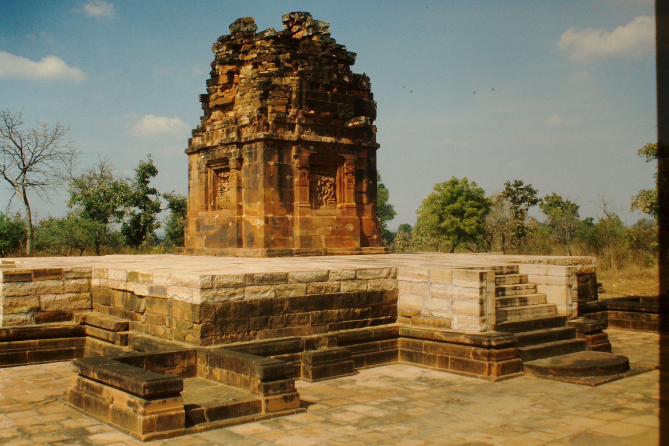 A square sandstone shrine with a ruined, stepped upper storey and carved relief niches stands on a wide, low stone terrace. Stepped plinth courses and a projecting staircase are in the foreground, under a blue sky with trees behind.