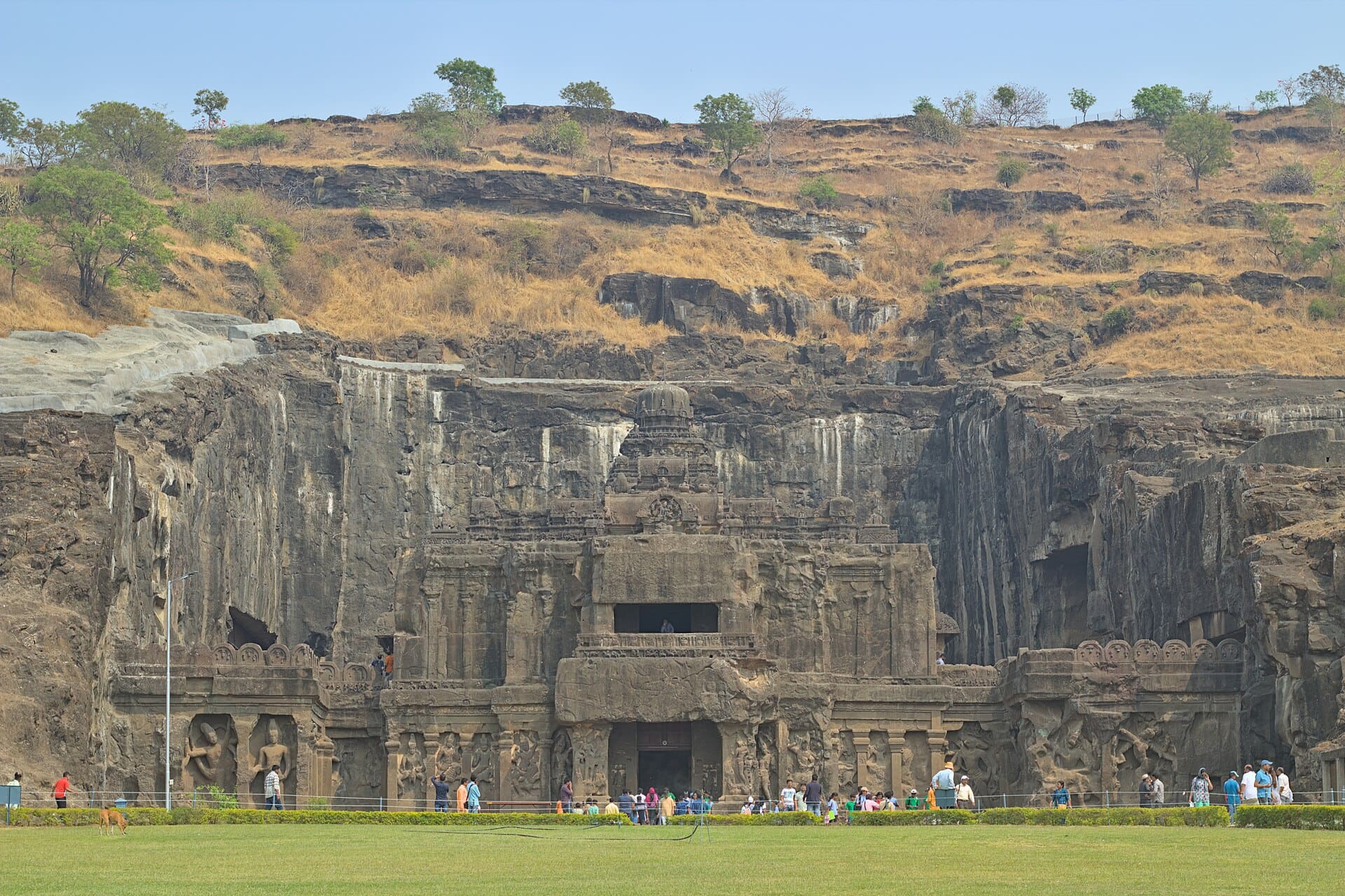 The rock-cut entrance façade of Kailāsa Temple at Ellora, photographed from the lawn outside. A wide wall of sculpted basalt with pillared bays, carved figures and a doorway, visitors along its base for scale, and the dry grass and rock of the escarpment rising above it.