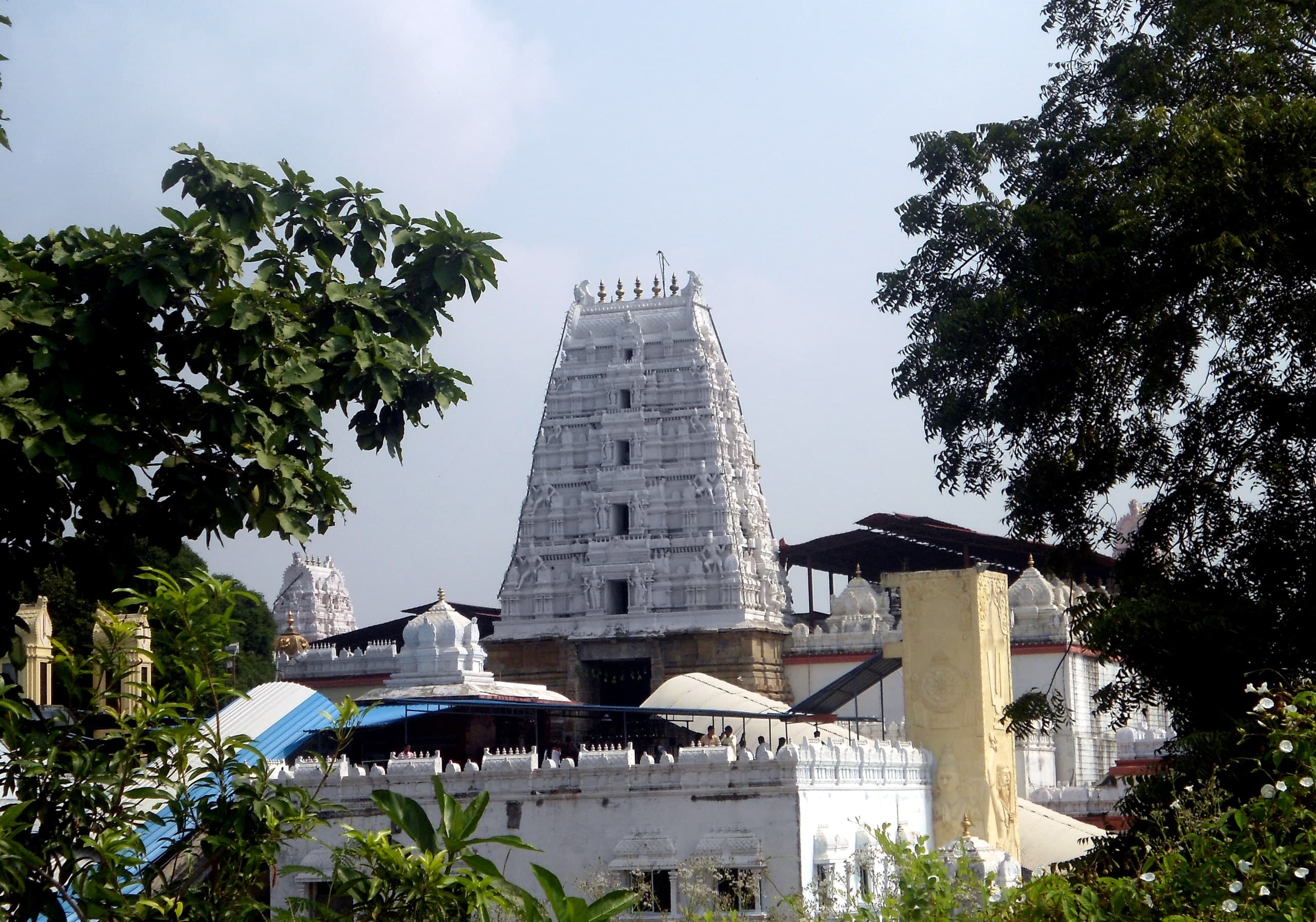 A white stepped gateway tower covered in small sculpted figures, rising above white enclosure walls and a curved metal canopy, framed by leafy branches under a pale sky.