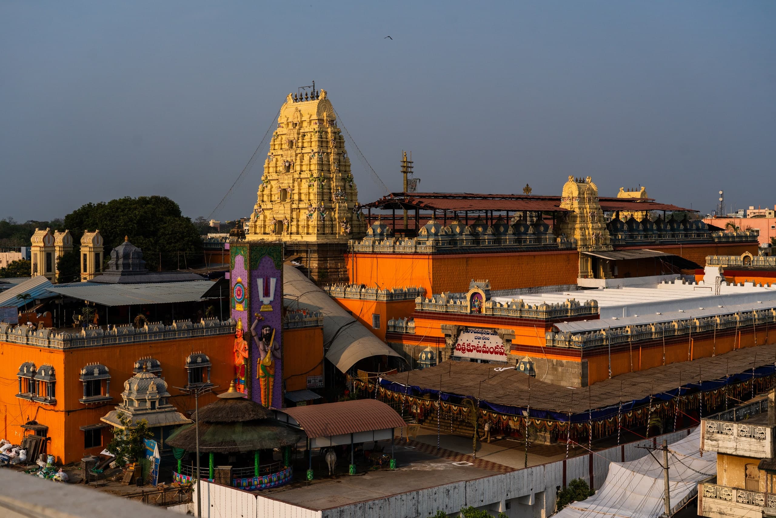 A tall cream-and-gold stepped gateway tower above orange enclosure walls in low evening sun. In front stand a tall purple panel painted with a Vaiṣṇava forehead mark and two standing figures, a thatched pavilion and a long canopied hall hung with garlands.