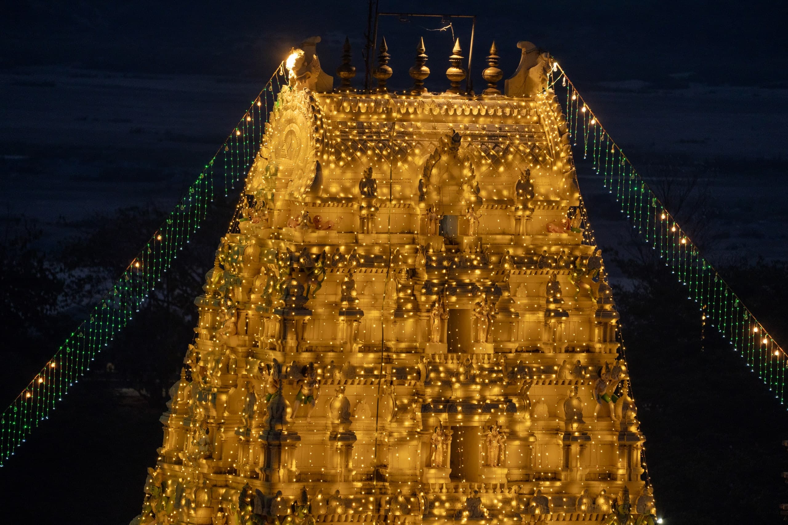 The upper tiers of a temple gateway tower glowing gold under hundreds of small electric lights, with strings of green and white lights running down from its top against a dark night sky.