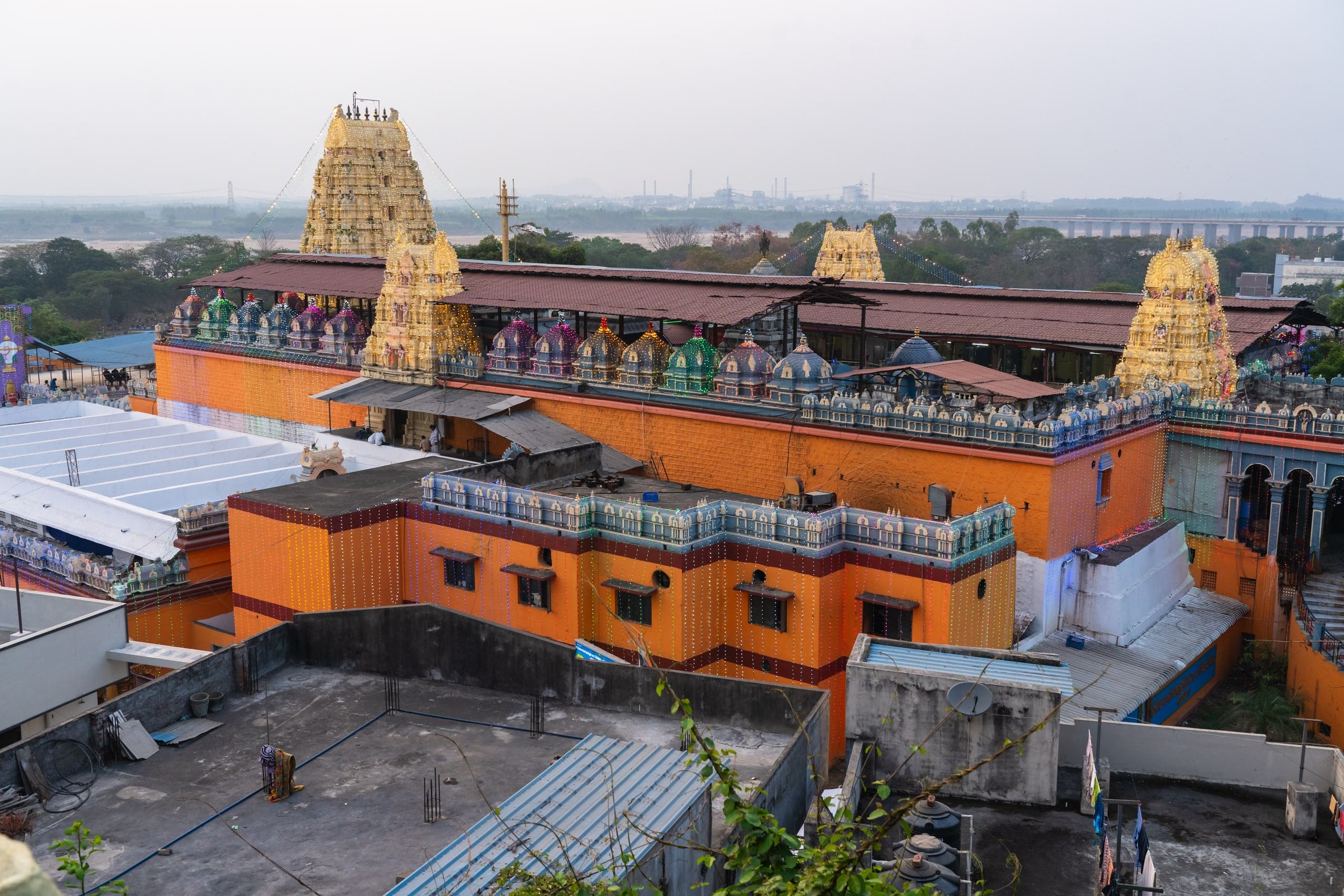 An elevated view over orange temple walls and four gold-painted gateway towers strung with lights, with a long roofed hall behind them, and a wide sandy riverbed, a long bridge and distant factory chimneys in the haze beyond.