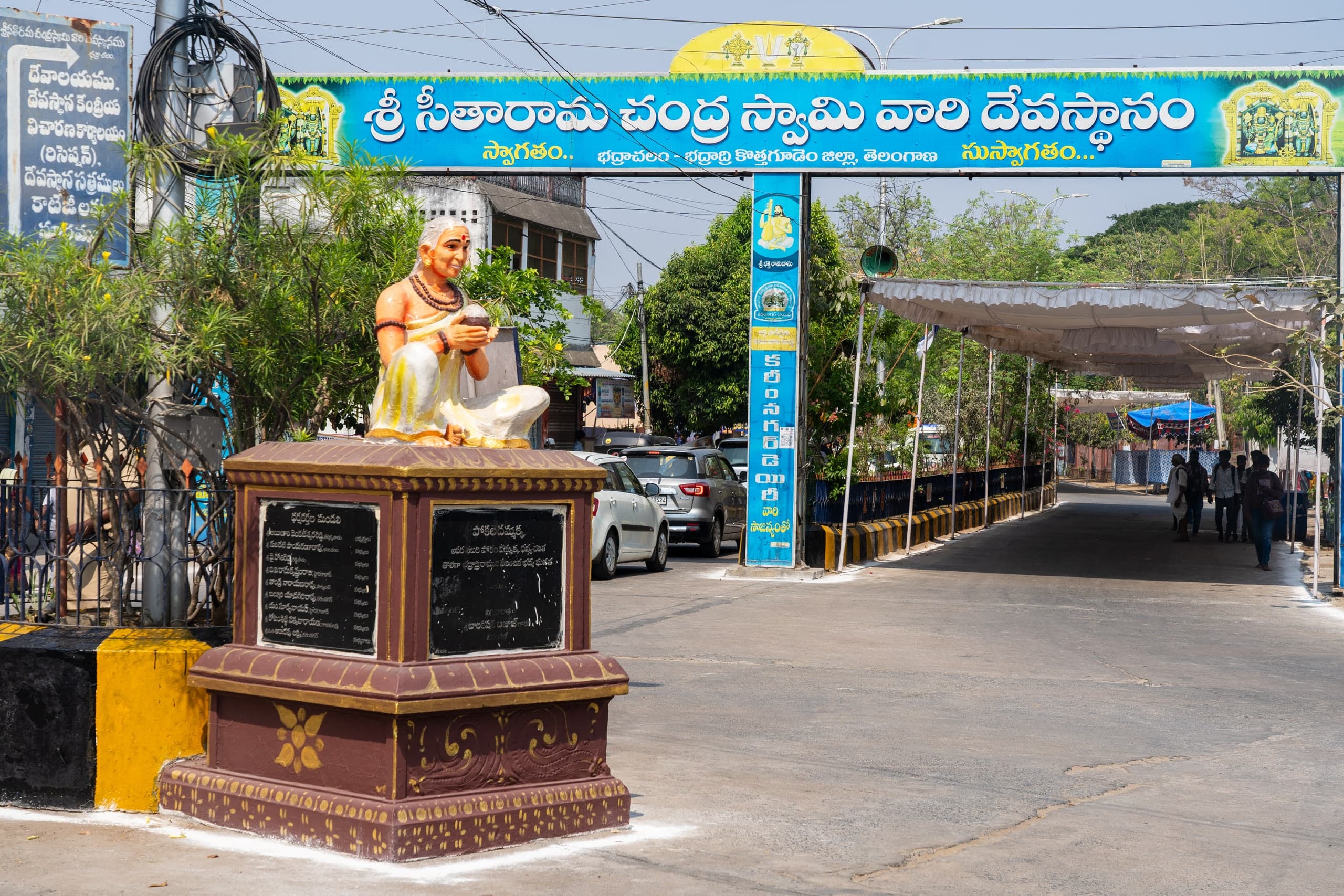 A brightly painted statue of a grey-haired woman seated cross-legged with a bowl in her hands, on a maroon pedestal with two black inscribed plaques, beside a blue welcome arch lettered in Telugu over a road with parked cars and a long white canopy.