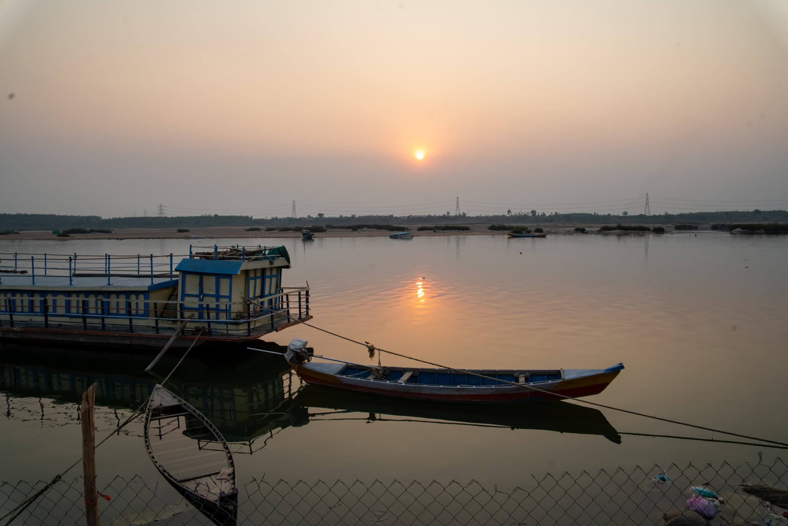 The sun low in a hazy sky over a wide, calm river, with a moored ferry with a blue and cream cabin and two wooden boats in the foreground, a wire fence along the bank, and power pylons on the flat far shore.