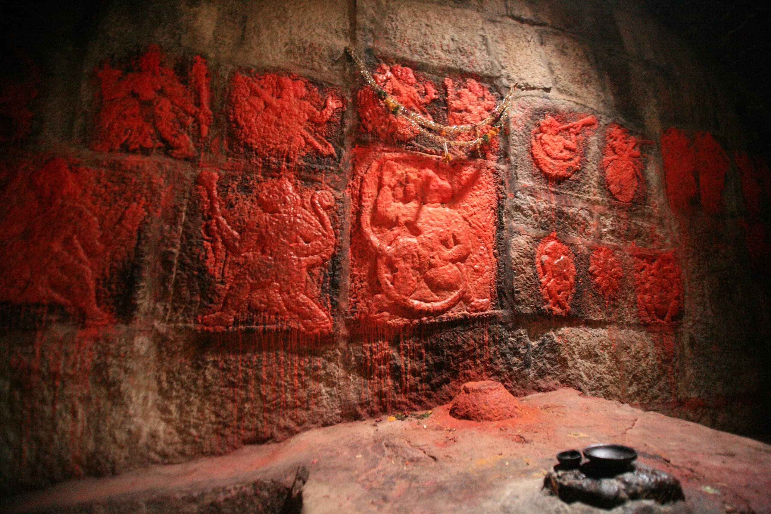 Rows of worn relief panels on a stone-block wall, thickly coated in red vermilion and hung with a tinsel garland, above a rock floor with a small heap of vermilion and two small black dishes.