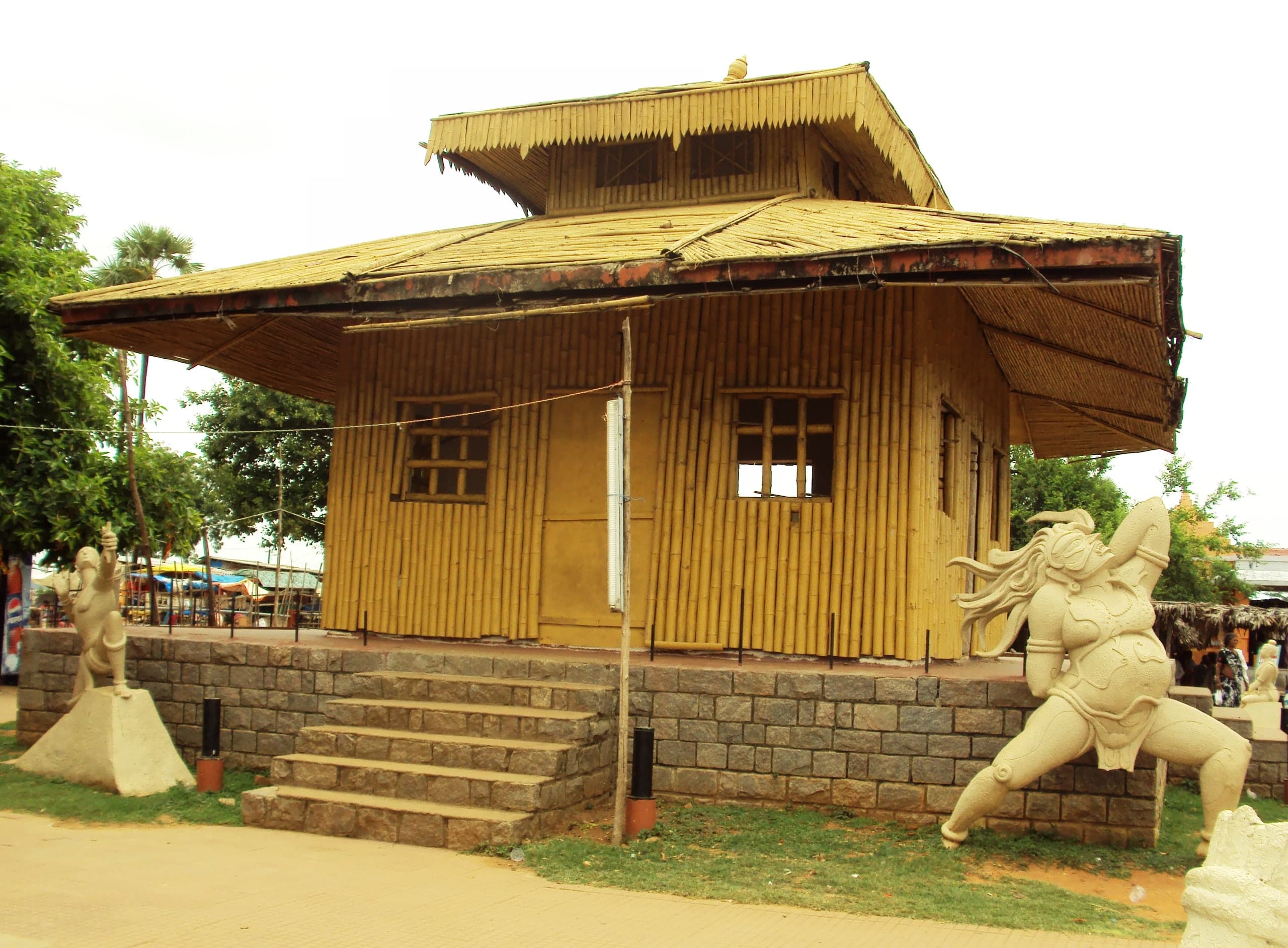 A bamboo hut with a two-tier thatched roof standing on a raised plinth of grey stone blocks with steps, flanked by two large pale cement statues: a slender figure with a raised arm on the left and a muscular figure with streaming hair on the right.