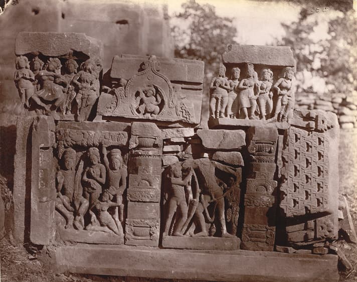 Sepia photograph of carved stone fragments in two tiers, the lower row set between carved pilasters on a base course, including panels of standing and dancing figures, an arched niche with a seated figure, and a lattice-patterned slab.