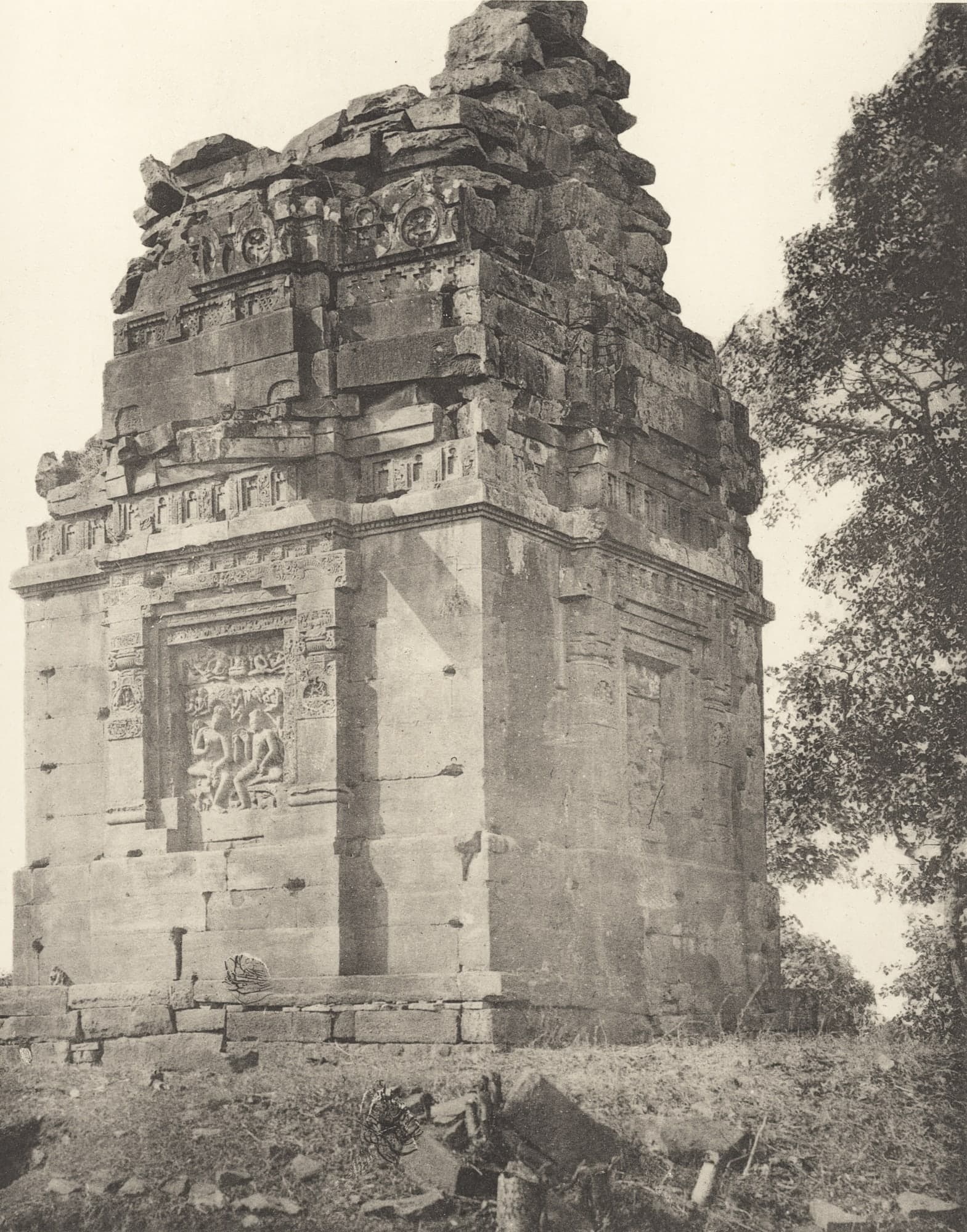 Black-and-white photograph of a square stone shrine with a ruined stepped top. A framed relief of two seated figures is set in one wall; a tree stands at right, and broken stones lie in the grass in front.