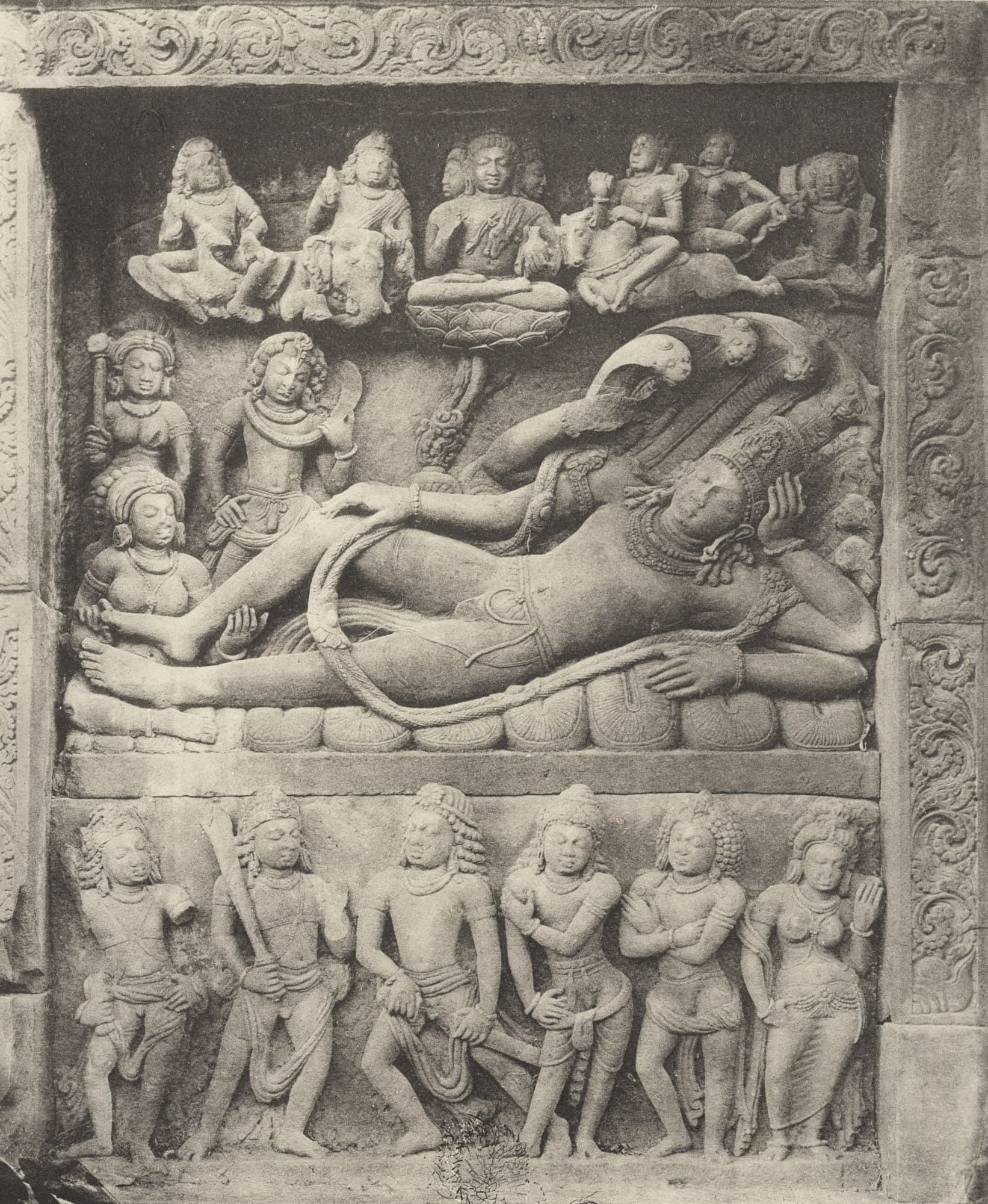 Black-and-white photograph of a framed relief: a crowned figure reclines on the coils of a many-hooded serpent while a seated woman holds his foot, with a row of seated figures above and six standing figures in a band below.