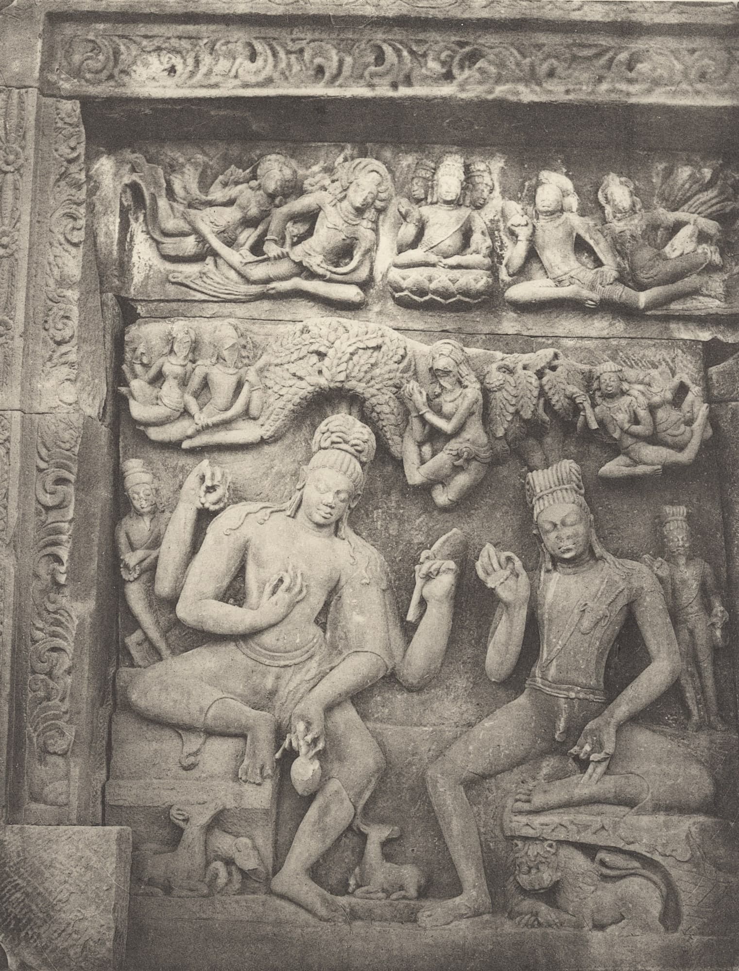 Black-and-white photograph of a framed relief of two seated ascetic figures with piled-up hair in a rocky setting, flying figures and a row of seated figures above them, and deer and a lion carved below.