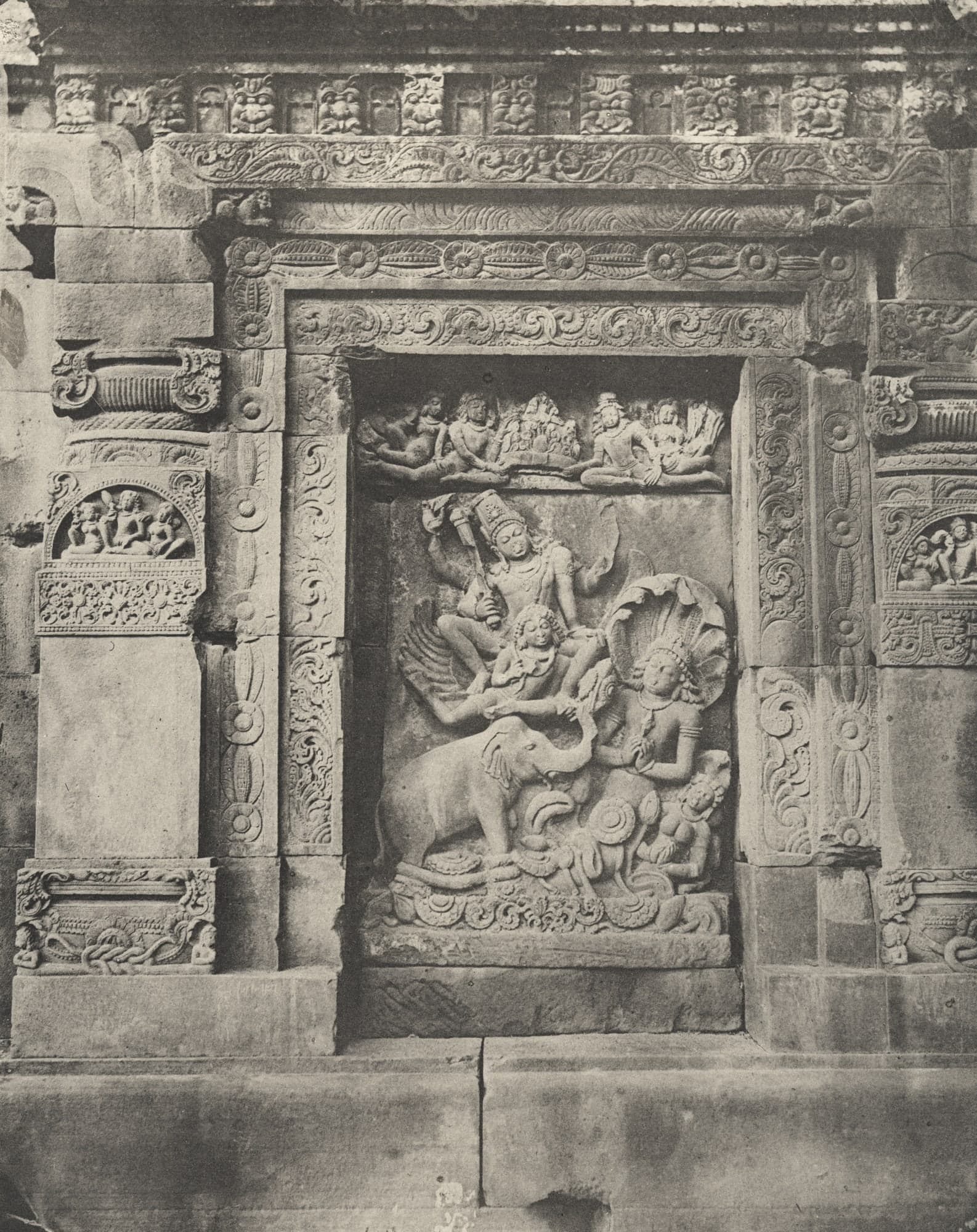 Black-and-white photograph of a framed relief niche: a crowned, four-armed figure rides a winged figure above an elephant whose raised trunk holds a flower, with a serpent-hooded figure beside it and a row of seated figures above.
