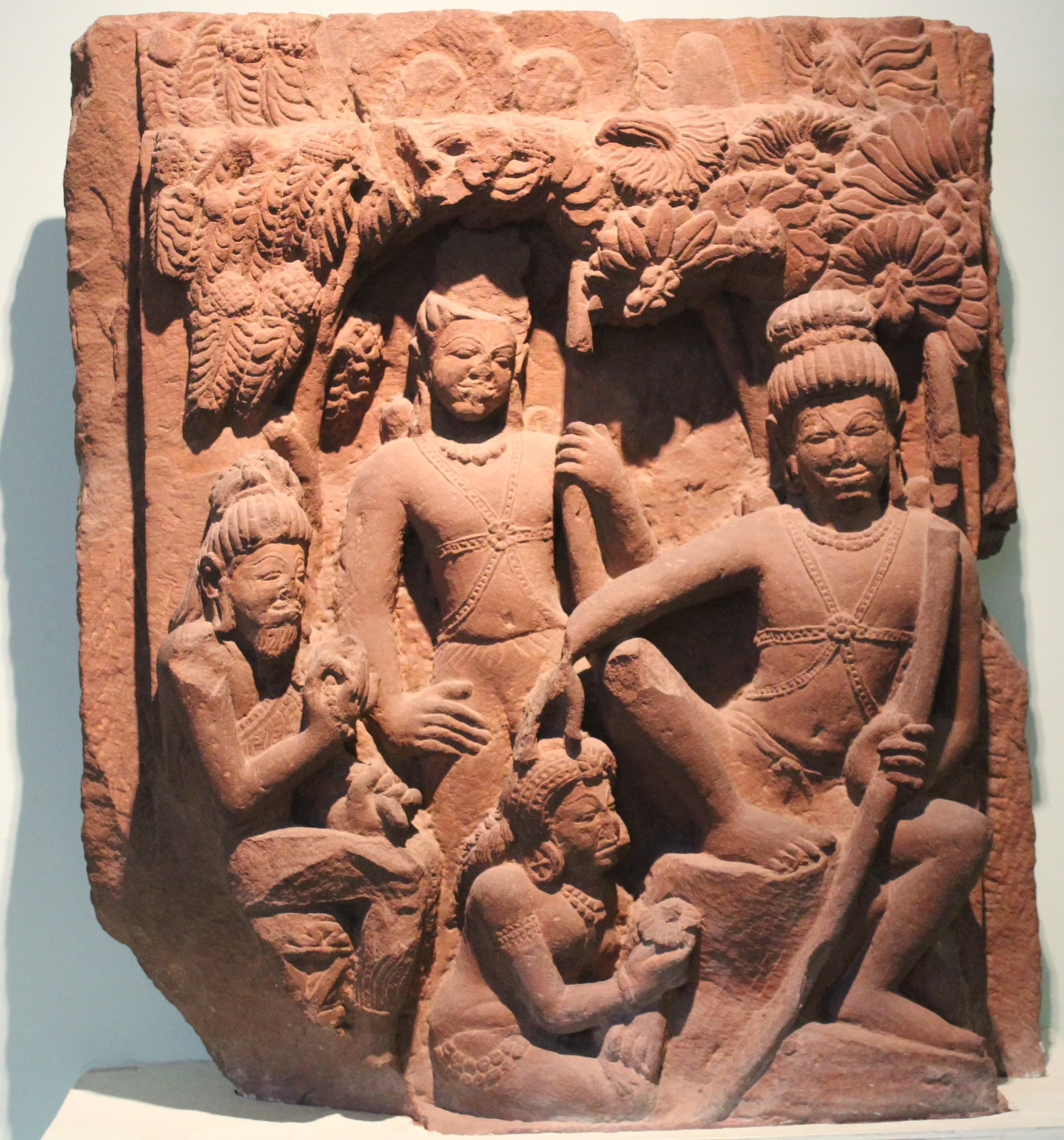 A red sandstone relief, its lower-left corner broken, under carved foliage: a bearded ascetic sits at left, a youth stands in the centre, a second youth sits at right holding a long bow, and a woman kneels between them.