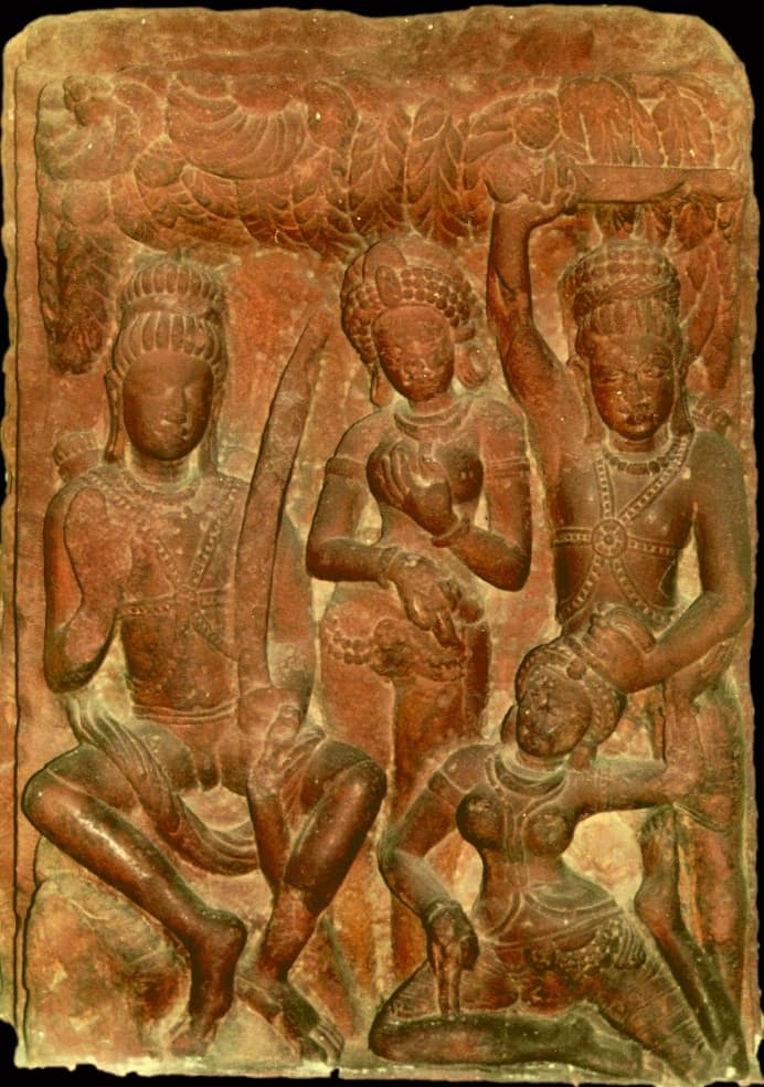 A red sandstone relief under a canopy of carved leaves: a seated man holding a bow at left, a standing woman in the centre, and at right a standing man with one arm raised, gripping the hair of a woman kneeling at his feet.