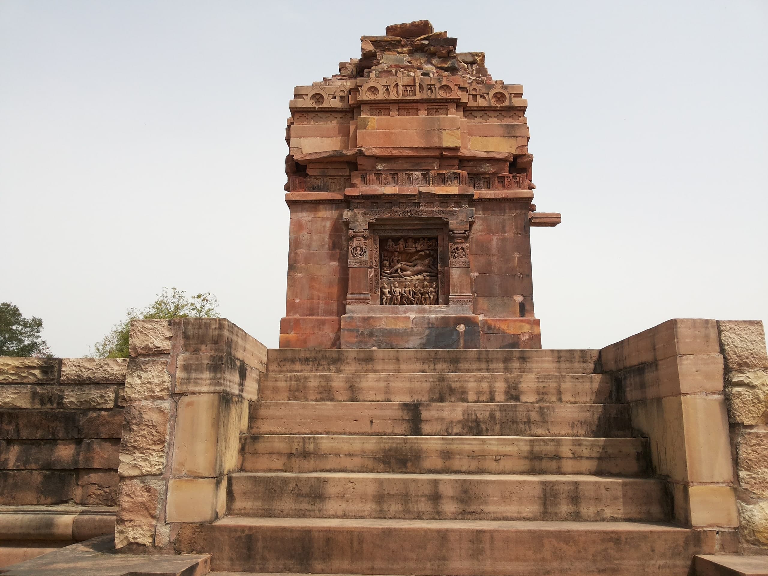 Broad stone steps lead up between low terrace walls to a square sandstone shrine with a broken stepped top. A framed relief niche on the wall facing the steps shows a figure lying on serpent coils above a row of standing figures.