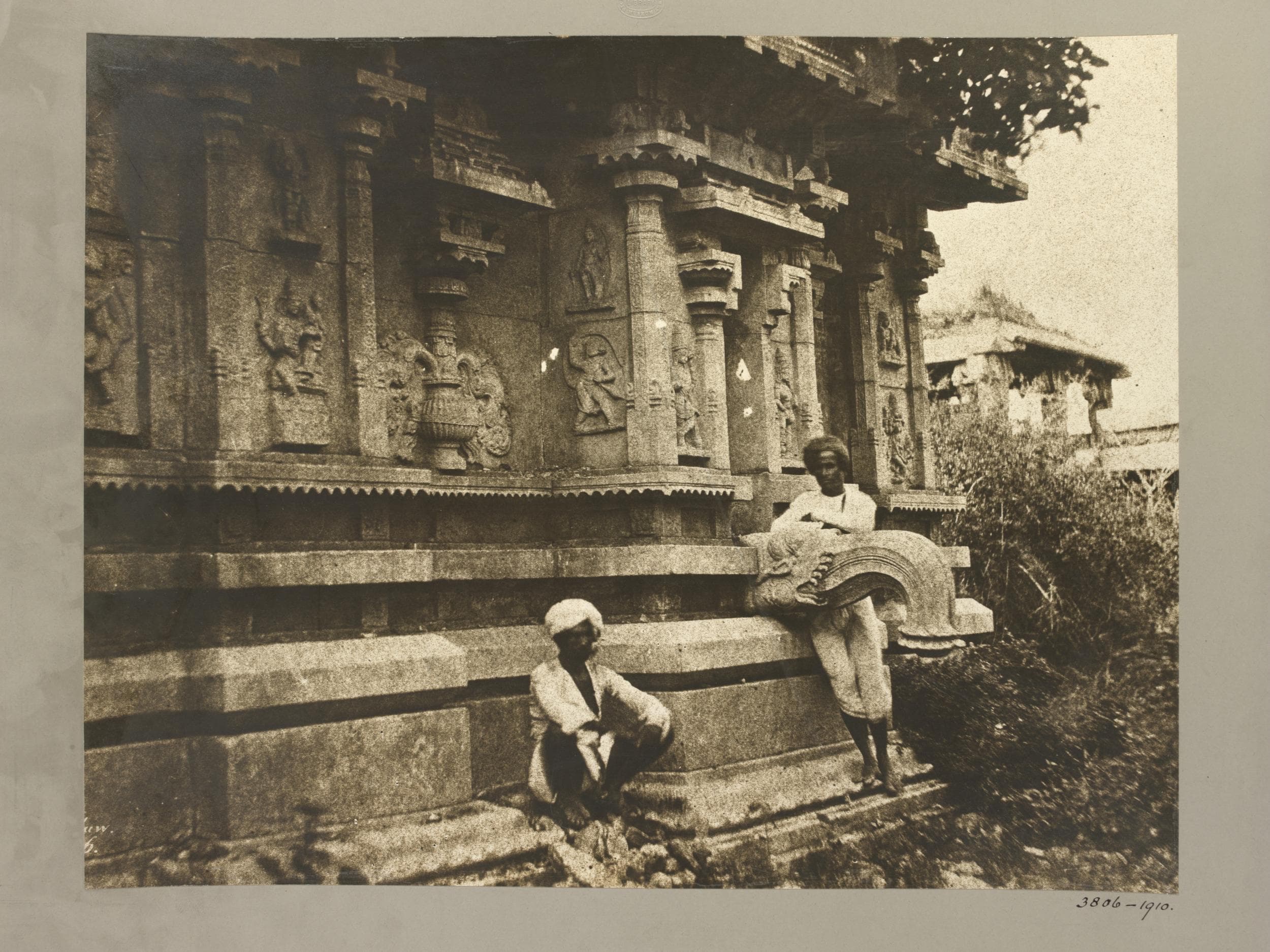 Sepia photograph mounted on grey card. A carved shrine wall with pilasters, niches of sculpted figures and a projecting roof rises from a moulded stone base. One man sits on the ground in front of the base; a second stands beside the curled stone balustrade of a stair. Shrubs and a ruined pavilion are at right.