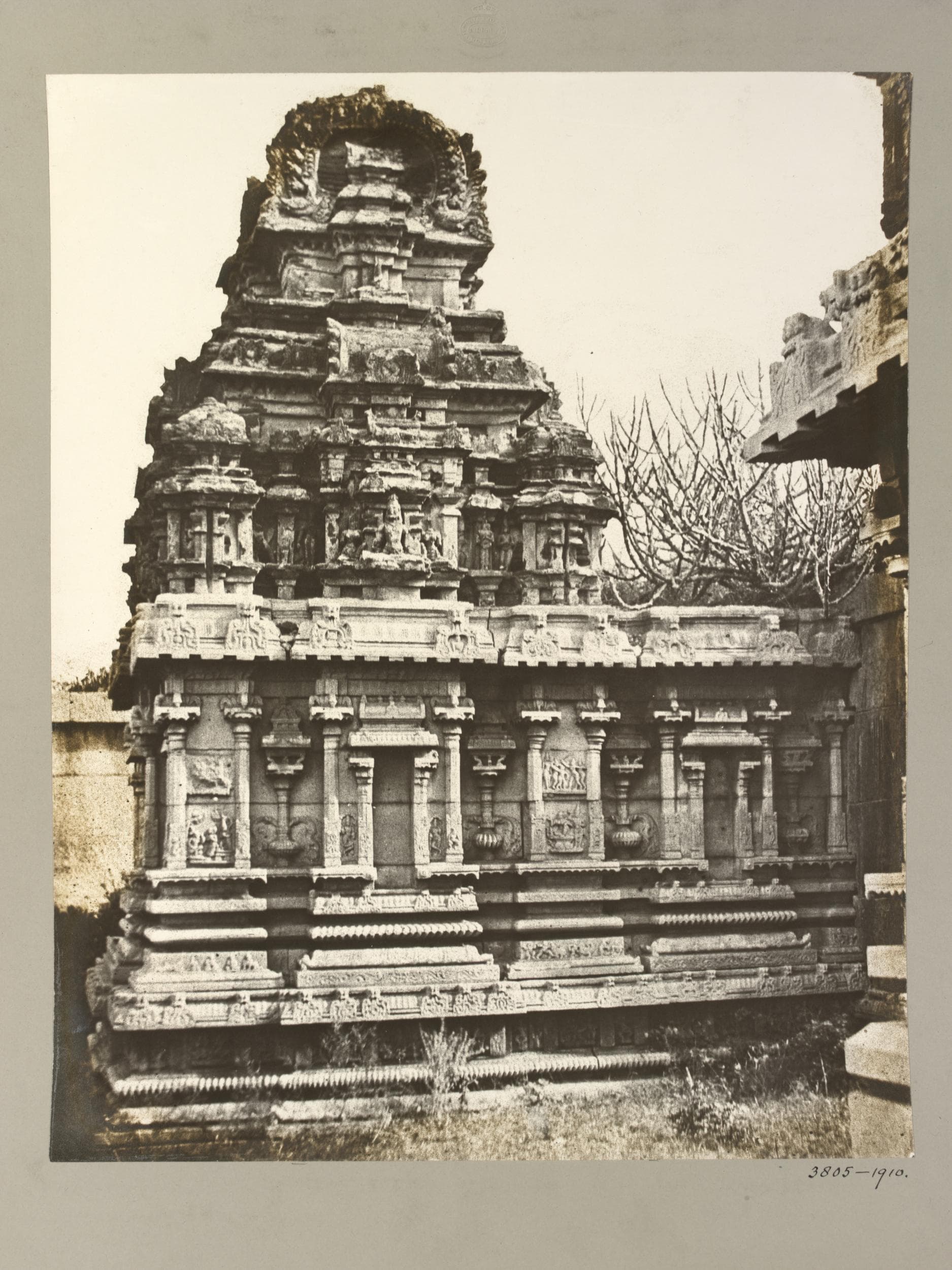 Sepia photograph mounted on grey card. A compact stone shrine on a moulded base, its walls divided by pilasters and niches, carries a stepped tower weathered at the top. Grass grows along the base and a bare tree stands behind.