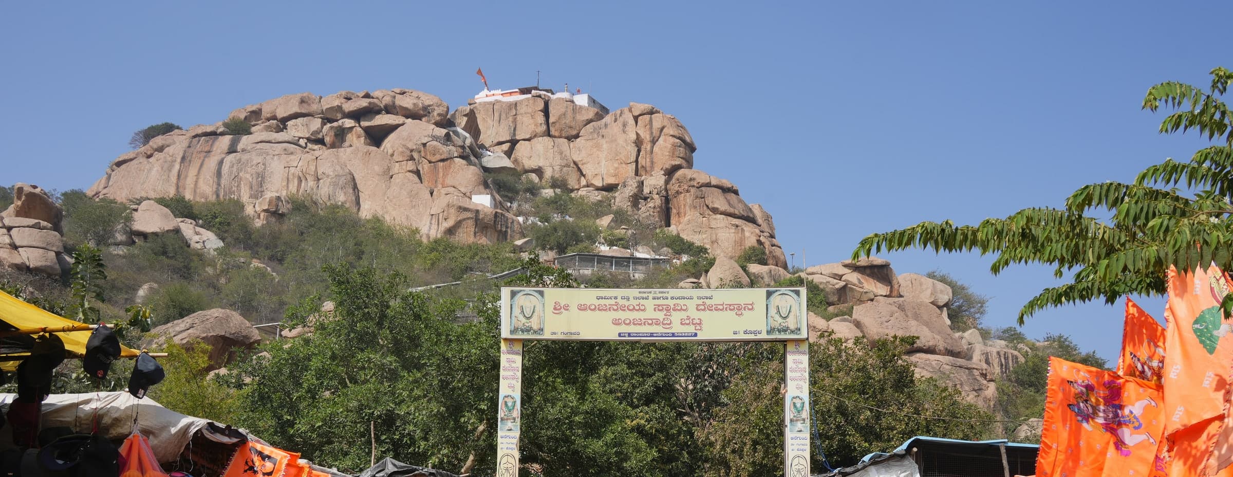 A rocky granite hill under a clear sky, topped by a small white shrine with a saffron flag. Below, a gateway arch with a Kannada inscription stands among trees, with saffron pennants and stall awnings at the edges.