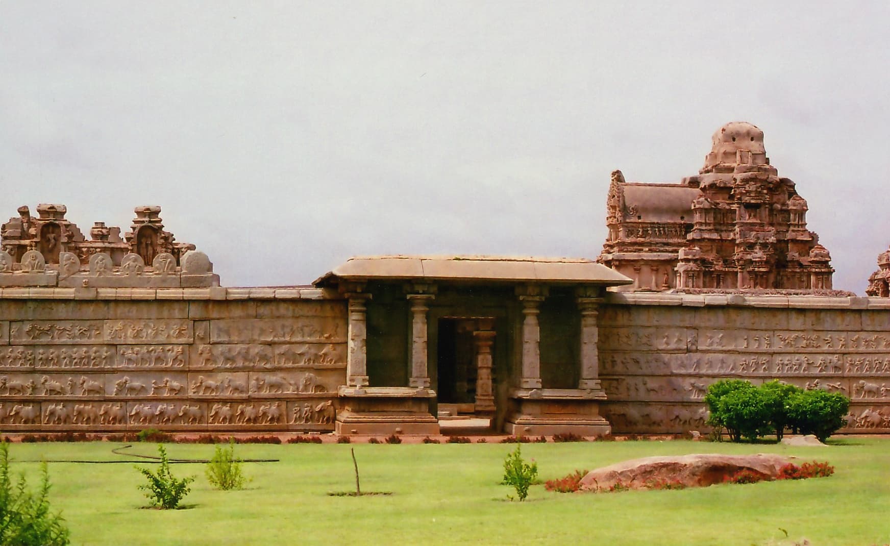 A long, low stone enclosure wall covered in horizontal bands of carved figures, with a pillared entrance pavilion at its centre. Behind the wall at right rises a stepped stone shrine tower; a lawn lies in front.