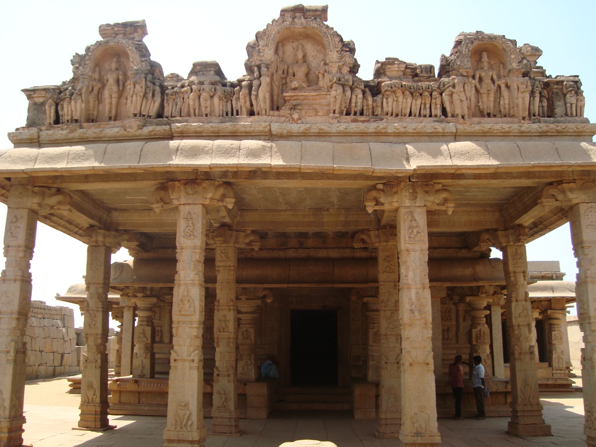 Frontal view of a pillared stone porch with a sculpted parapet of three arched niches and rows of standing figures. Through the pillars, a dark doorway opens into an inner hall. Two visitors stand at the right.