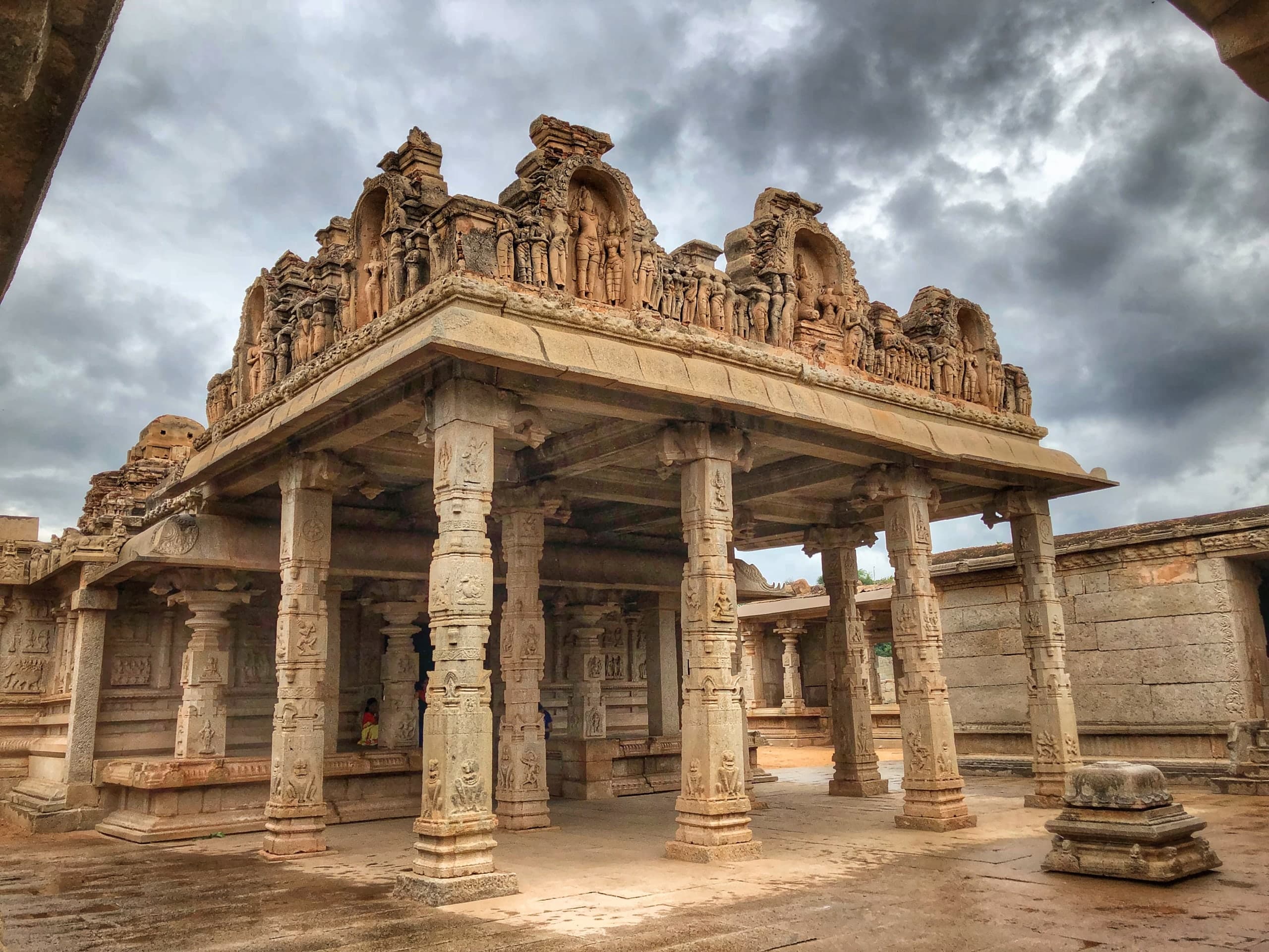 A granite pillared porch under a stormy sky. Slender carved pillars carry a flat stone roof topped by a parapet of sculpted niches crowded with standing figures. Behind it rise the walls of the enclosure; a carved pedestal stands on the wet paving in the foreground.
