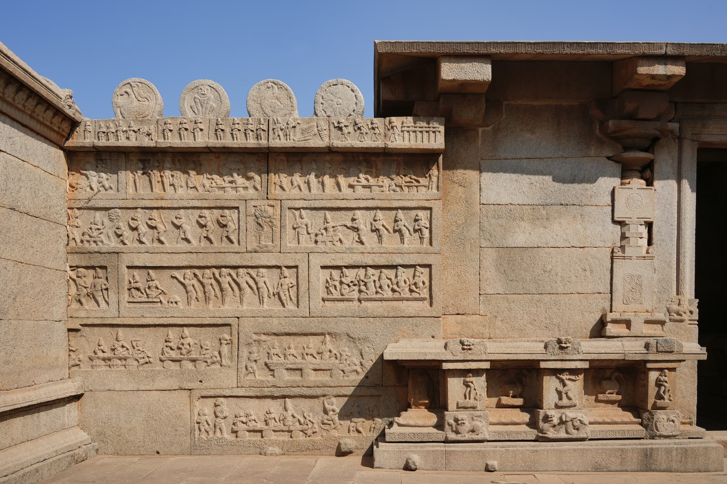 A stone wall carved with several horizontal rows of small figurative scenes, standing and seated groups, beneath a row of carved roundels against a blue sky. At right, a pillared pavilion with a carved base.