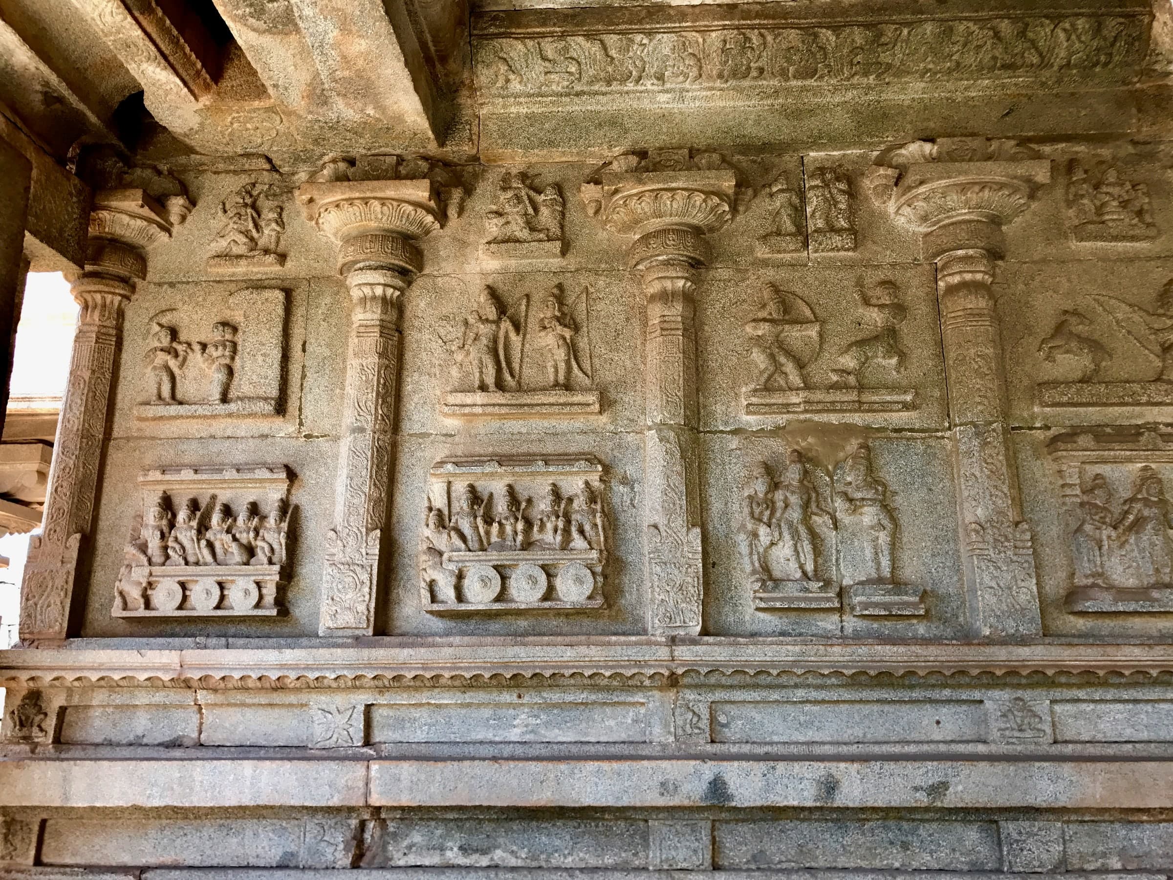 A grey granite wall under a projecting eave, divided by carved pilasters into two tiers of framed relief panels. In the lower tier, wheeled chariots carry rows of seated figures; above, standing figures and an archer drawing a bow.