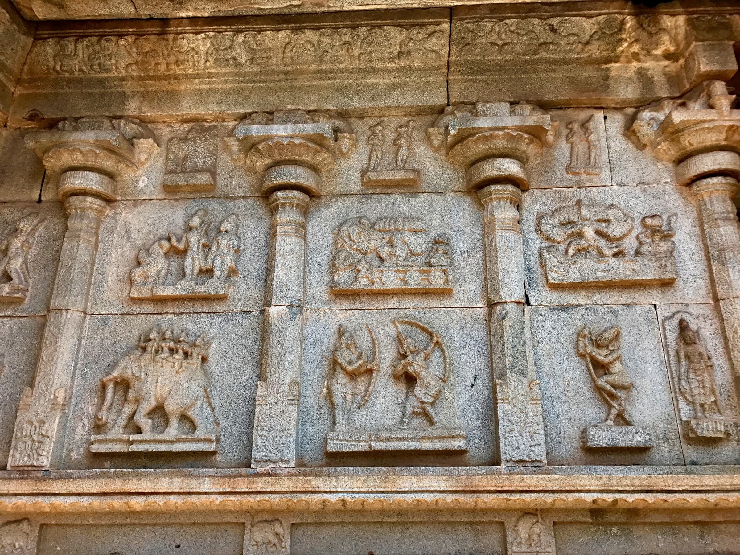 Two tiers of framed relief panels between pilasters on a sunlit granite wall: an elephant carrying several riders, two archers facing each other with drawn bows, and smaller scenes of standing figures above.