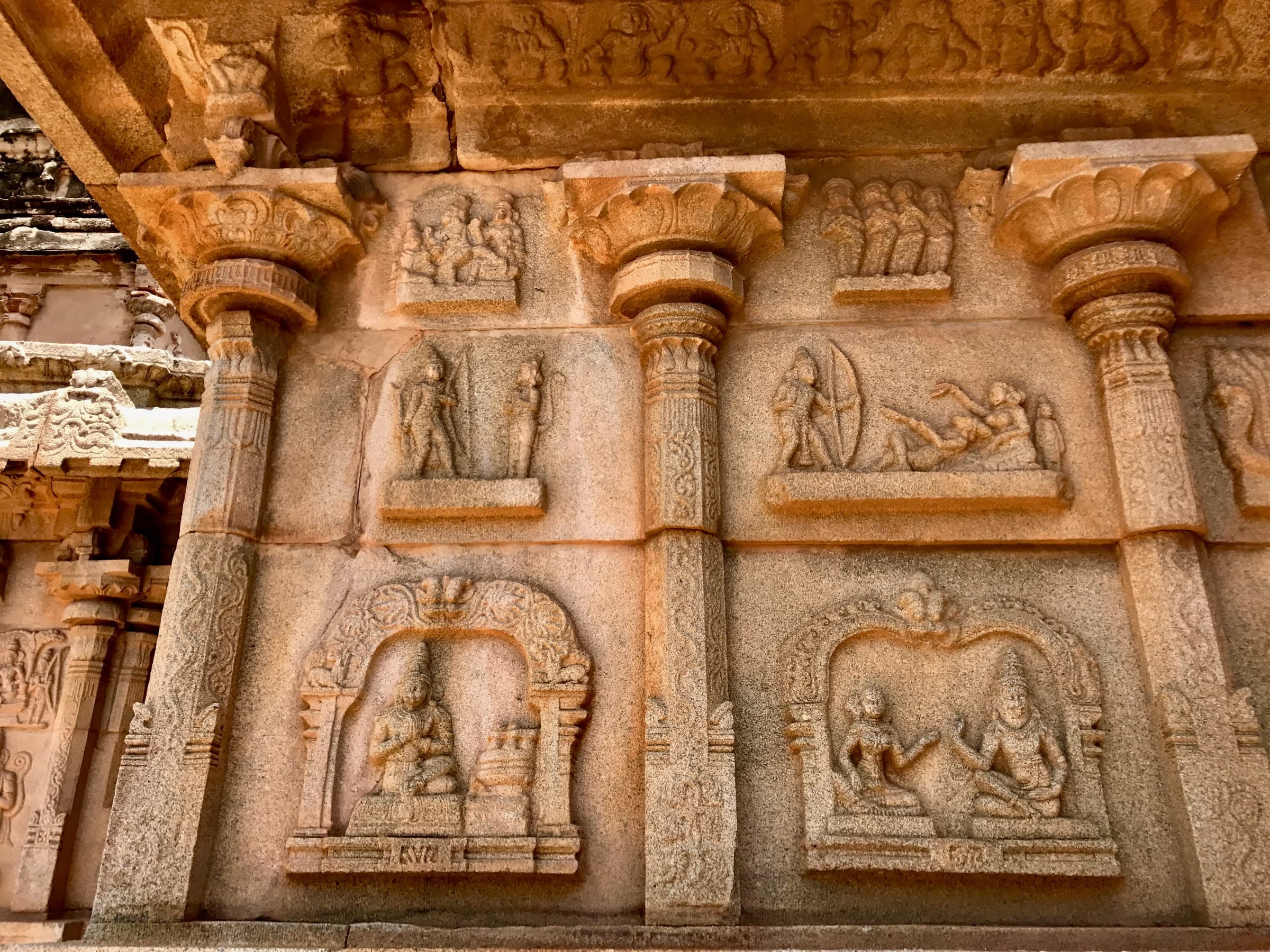 Warm-toned granite wall with carved pilasters framing relief panels: an archer and a reclining figure in the upper tier, and below, a seated figure in one ornate arched frame and a pair of seated figures in the other.