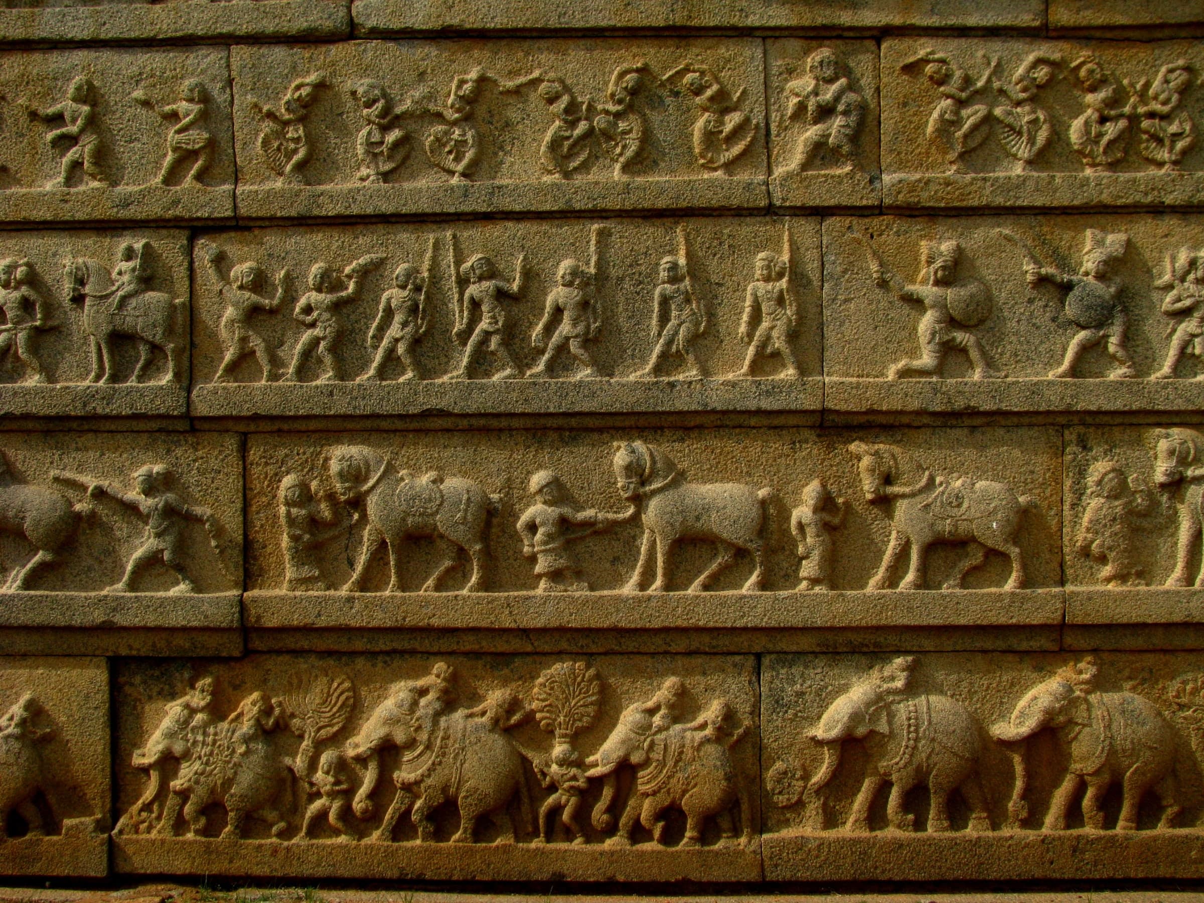 Close view of four stacked bands of relief on a granite wall. Bottom: elephants with riders and attendants. Above: horses led by grooms. Above that: men carrying spears, swords and round shields. Top: dancing figures with raised arms.