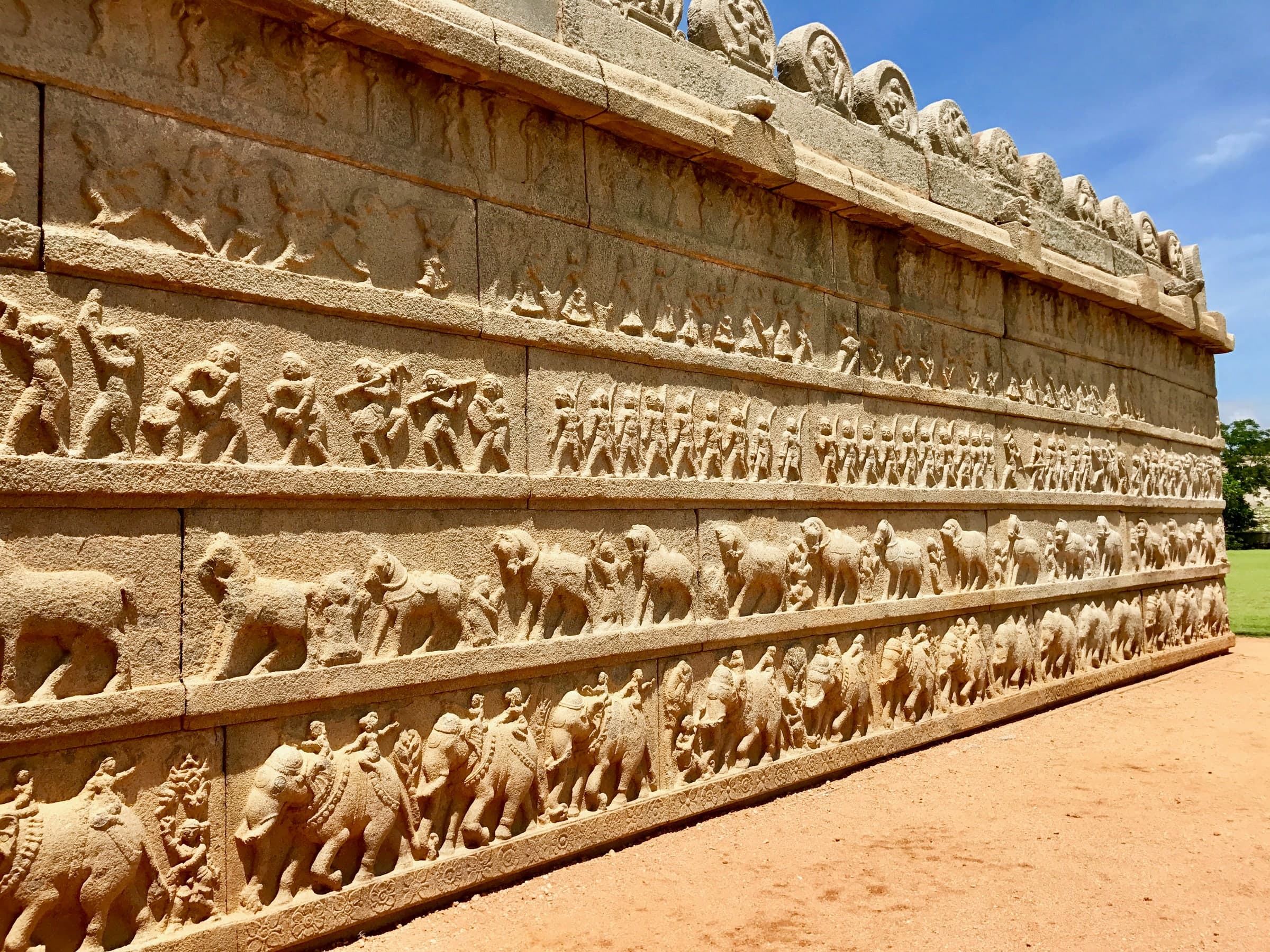 A long granite wall receding into the distance, carved in horizontal bands: a row of elephants along the bottom, a row of horses above, then rows of marching and dancing human figures, under a coping of carved roundels.