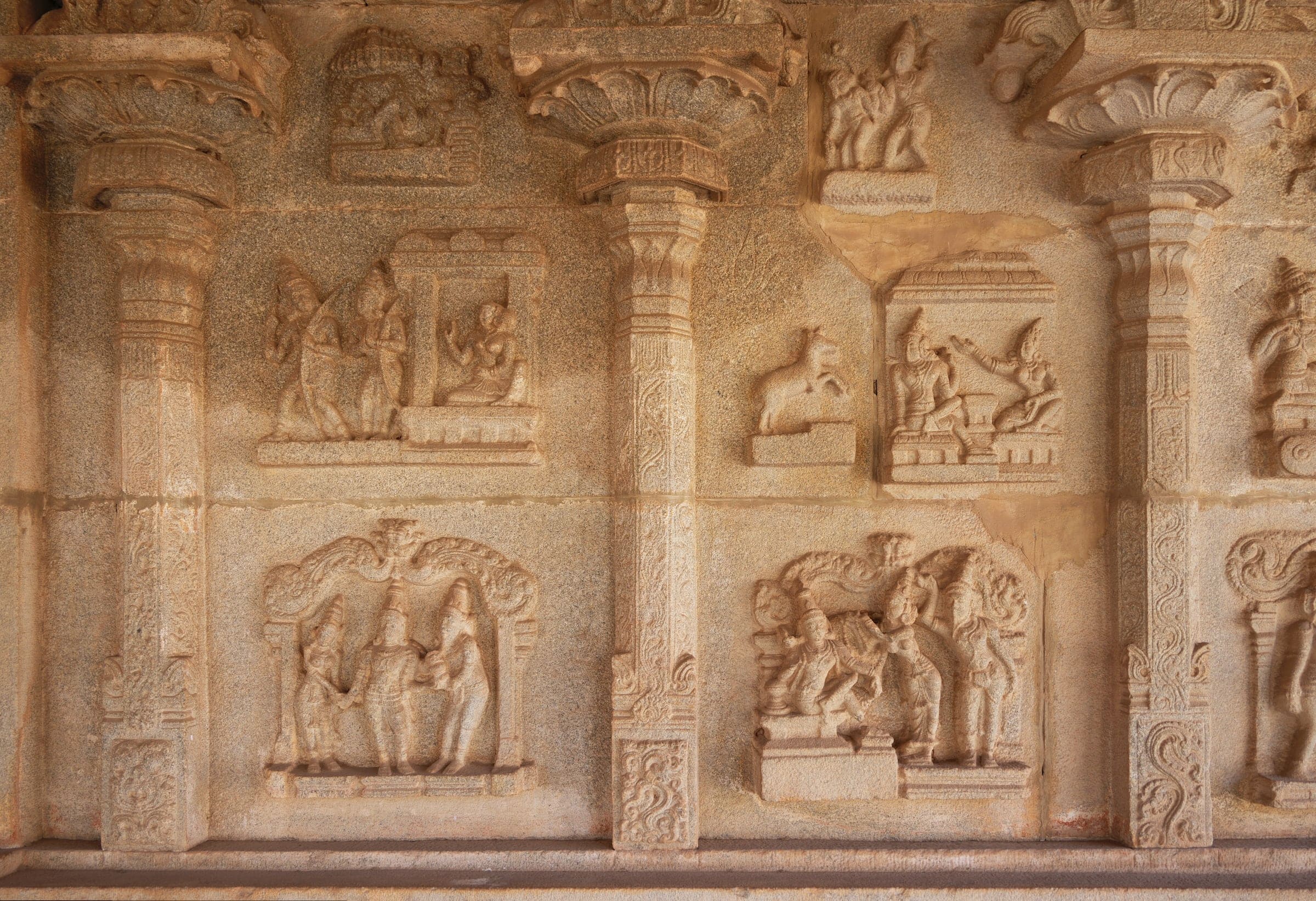 A warm-toned granite wall divided by tall carved pilasters into panels of relief. Small framed scenes show figures standing, seated and gesturing; one panel shows a lone deer. Some panels are worn or patched.