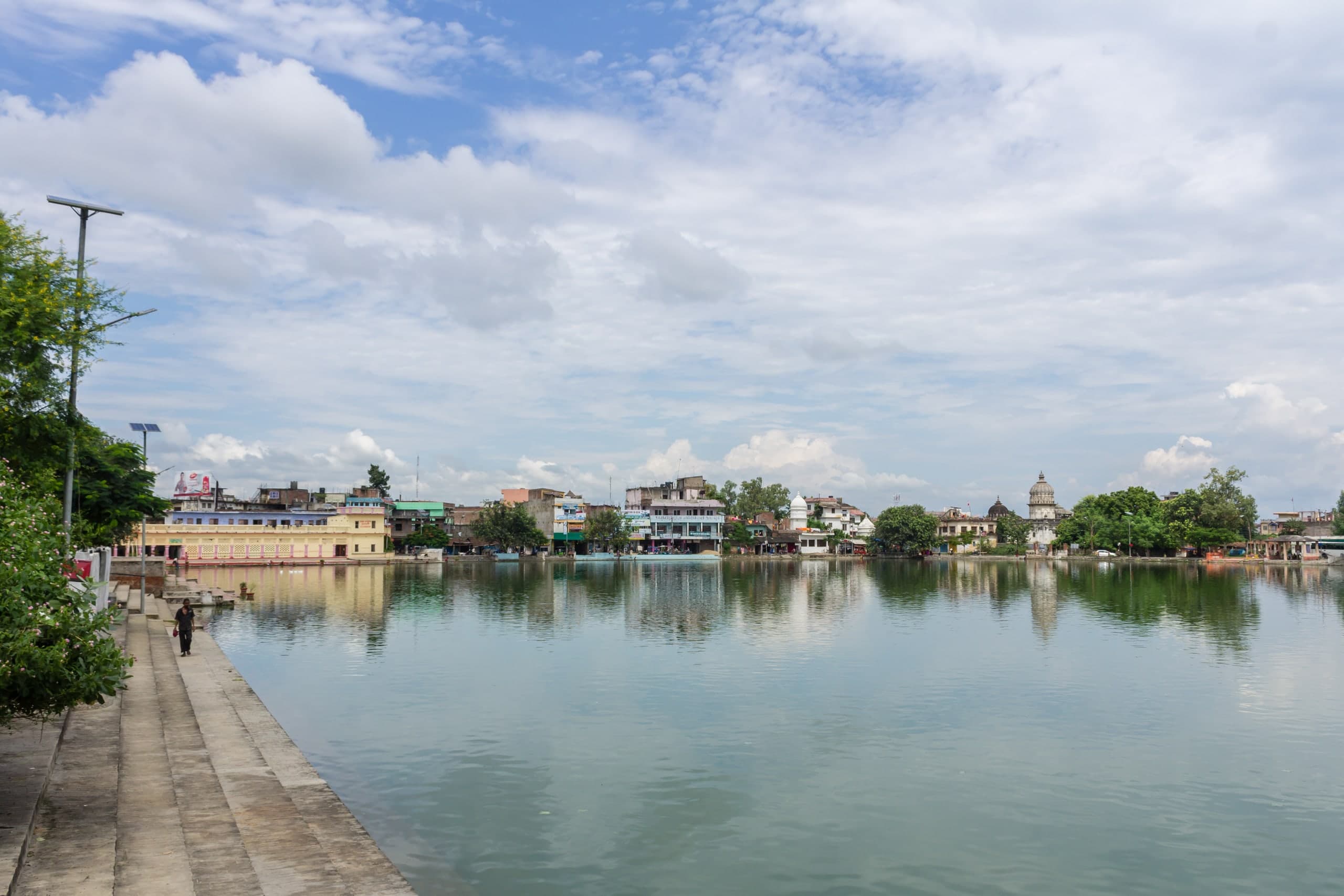 A large still pond reflecting a cloudy sky, with stone steps along its left bank and a line of town buildings and a white dome across the water.