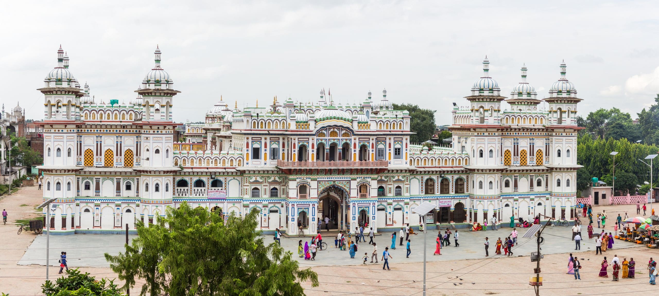 A long white palace-like building with blue, green and yellow painted trim, rows of arched windows and clusters of domed towers at each end, behind a paved forecourt where people walk.