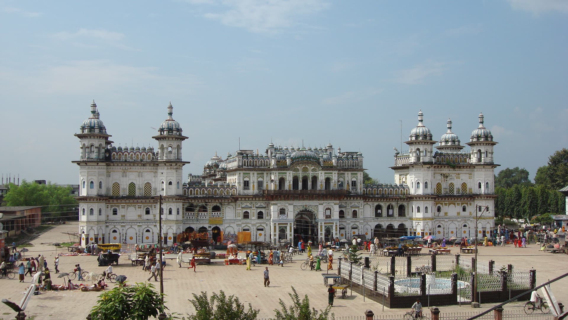 A white palace-like temple with domed corner towers seen across a busy open square with stalls, cyclists and a fenced fountain in front of it.