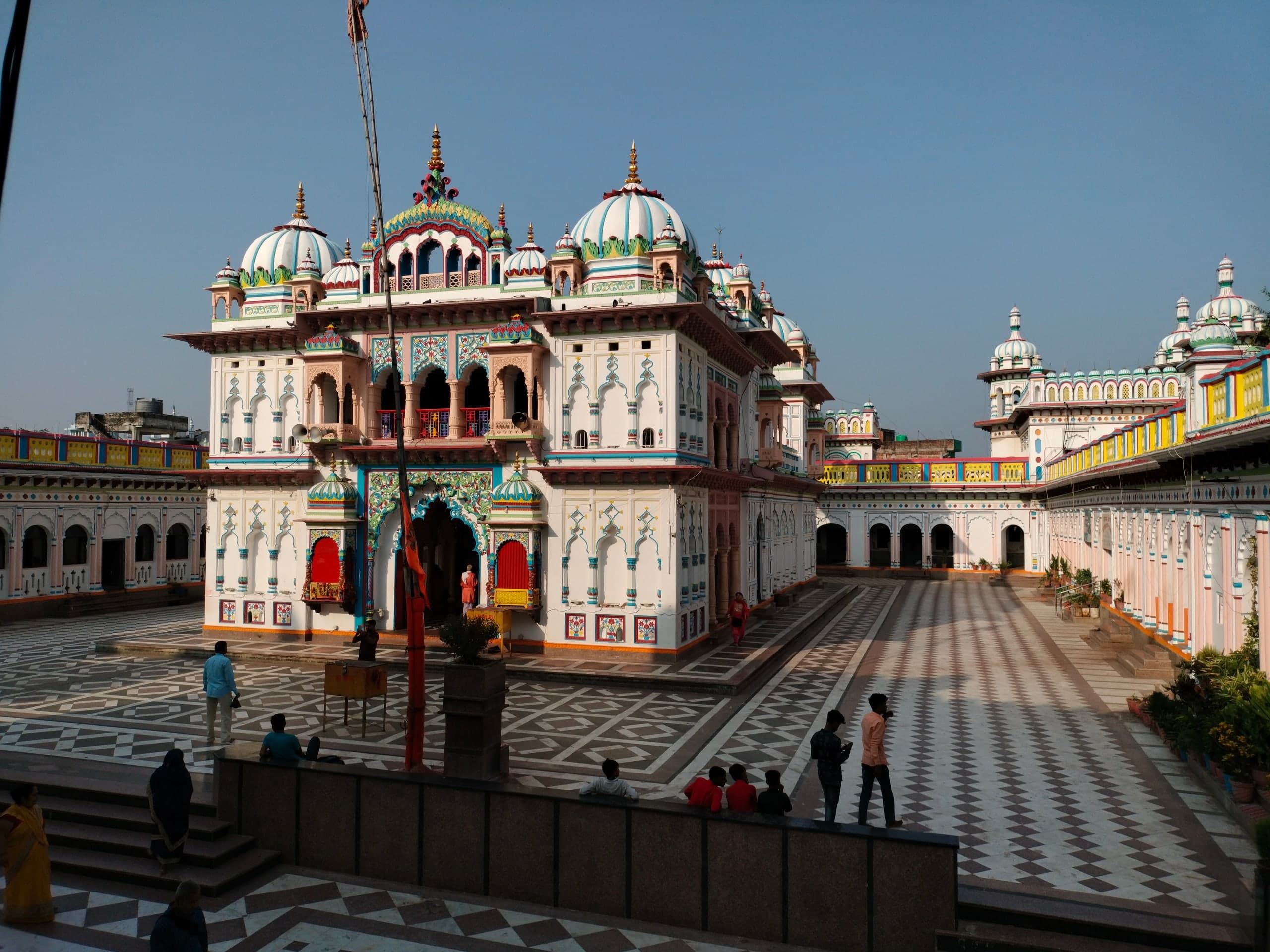 The front of a white domed shrine with a painted arched doorway, seen in late sun across a patterned courtyard where a few people stand by a low wall.