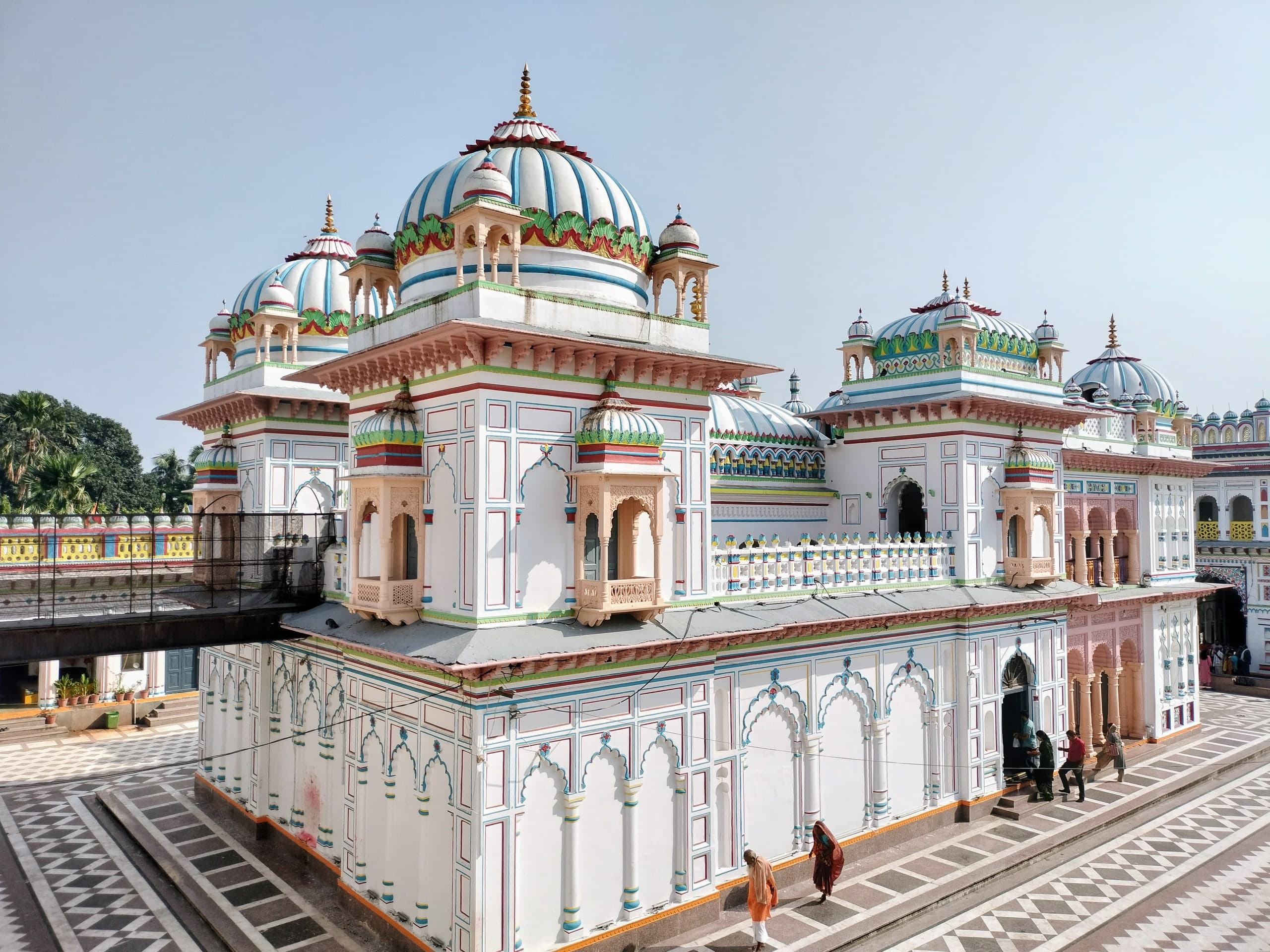 A white shrine with blue-striped domes and small domed balconies, its walls marked with painted arch outlines, standing in a courtyard paved in black and white patterns.