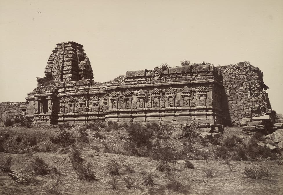 A faded sepia photograph of a long carved stone temple with a stepped tower at its left end. A rough rubble wall joins it on the right, and scrubby ground fills the foreground.