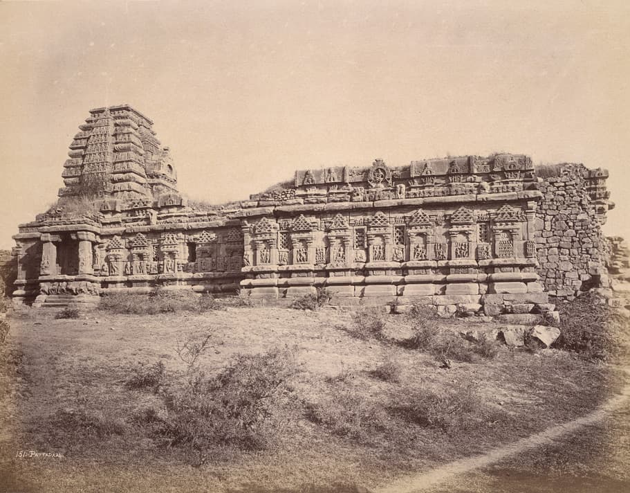 A sepia photograph of a long, low temple with rows of carved niches along its wall, a stepped curving tower at the left end, a rubble wall at the right and open, scrubby ground in front.