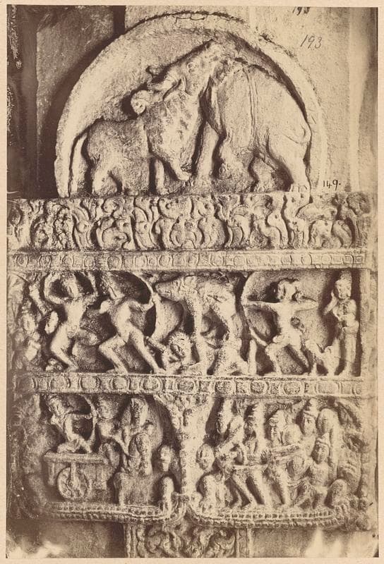 A sepia photograph of a carved column face. An arched panel at the top shows an elephant and a bull sharing one head, above two bands crowded with small figures, among them archers and a chariot wheel.