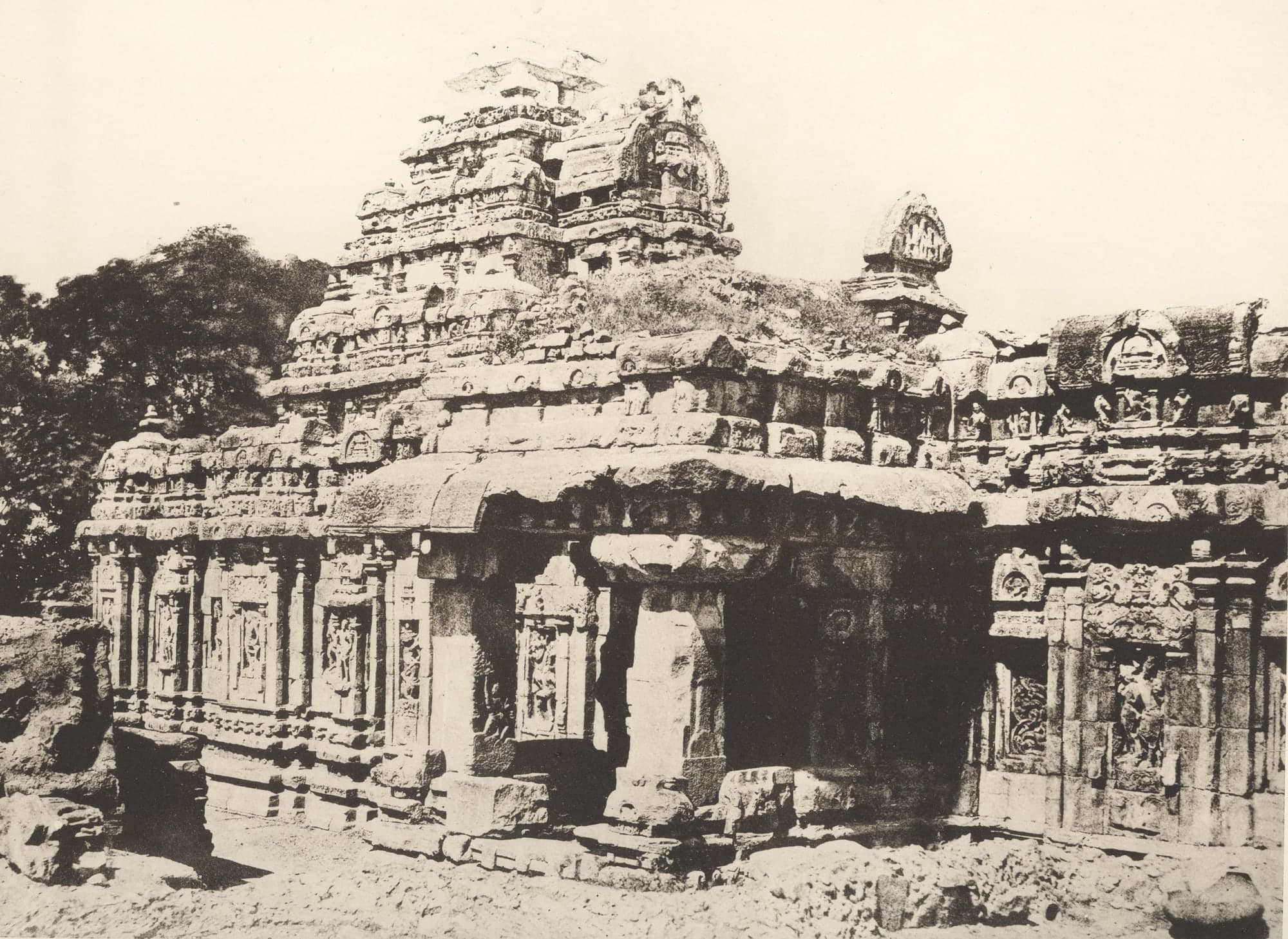 A sepia photograph of a carved stone temple with a deep, shadowed pillared porch, a tiered tower overgrown with grass above it, a small roof pavilion at the right and broken stones on the ground in front.